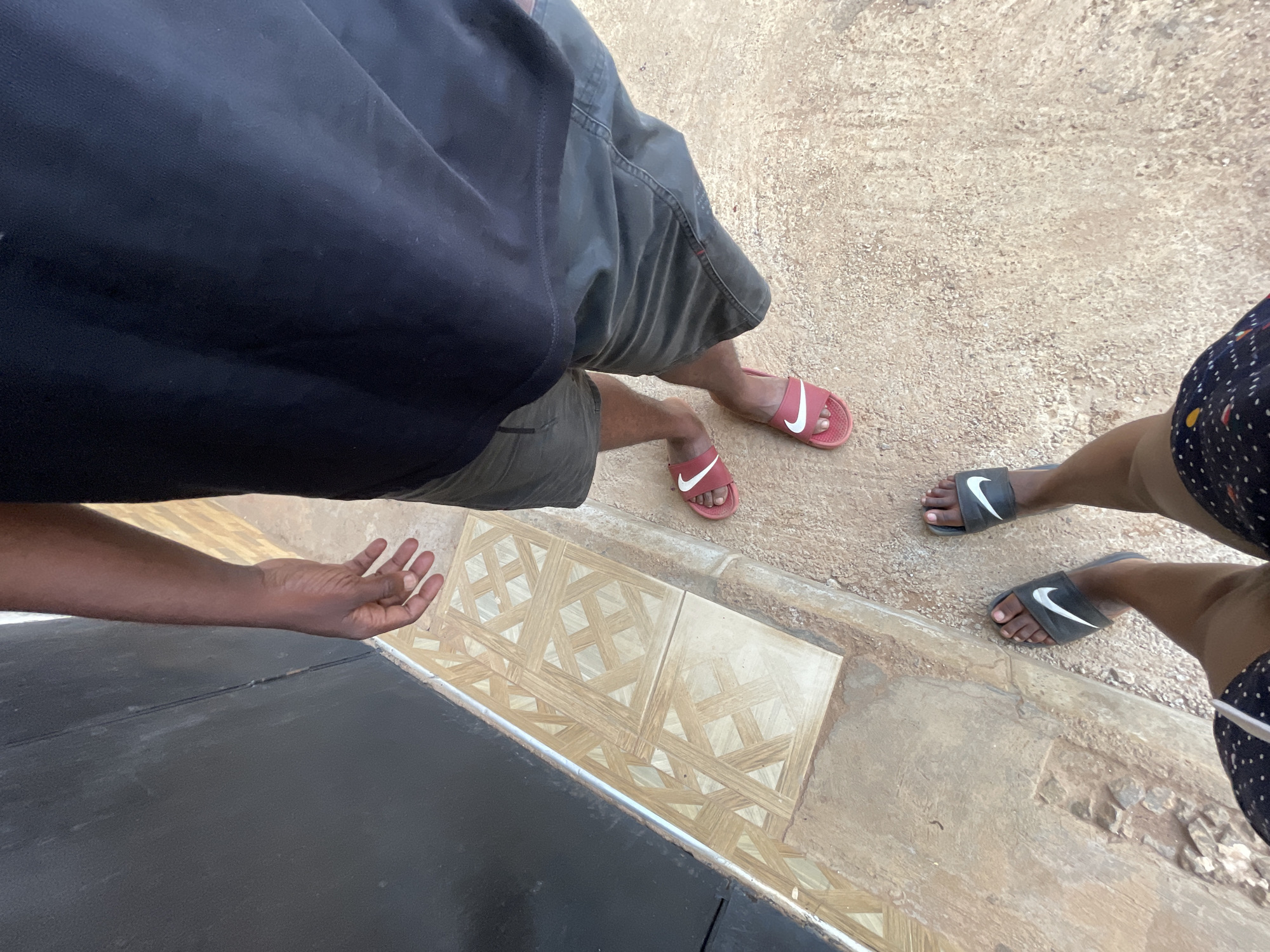 The image depicts a view looking down at two individuals standing near an edge, likely a sidewalk or curb. The person on the left is wearing a dark-colored top, dark shorts, and red Nike slides. Their arm is visible, with an open hand gesture. The person on the right is wearing a patterned dark garment, with black Nike slides on their feet. The ground is mostly a light brown concrete texture. There's an area of decorative tiles at the edge. The location is Kurunduma, Nigeria, as indicated in the prompt.