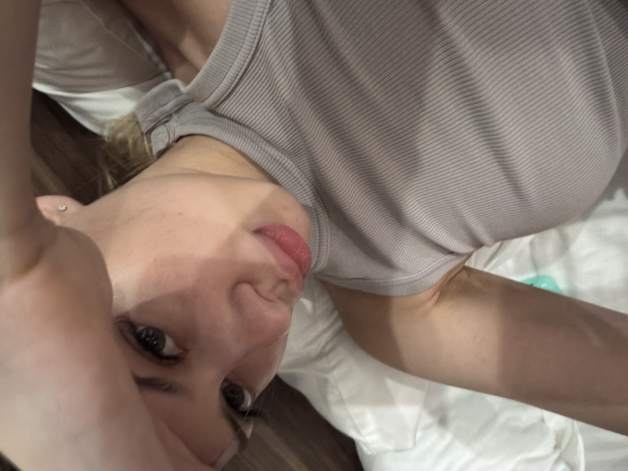 The image is a close-up selfie of a young woman lying on a bed. She is wearing a grey ribbed tank top and has her hand raised to her forehead. Her lips are slightly parted and her eyes are looking towards the camera. The background consists of white bedding and a brown wooden headboard. The lighting suggests an indoor setting, possibly a bedroom. There is no discernible information in the image to indicate a specific location within the city of Đà Nẵng, Vietnam.