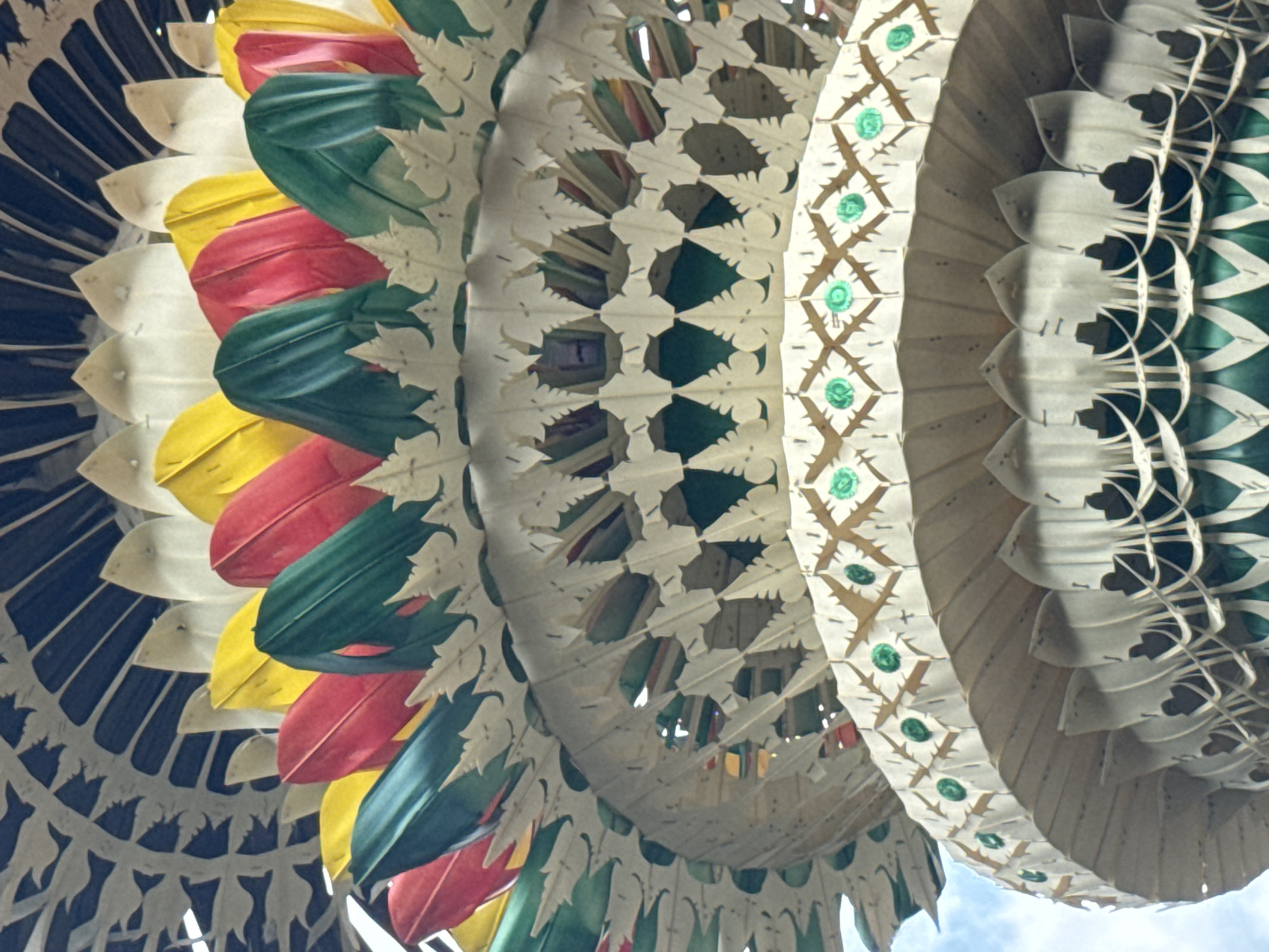 This is a close-up, low-angle, vertical shot of the top of a traditional Balinese *penjor*, a ceremonial bamboo pole, in Tegallalang, Indonesia. The *penjor* is intricately decorated with woven palm leaves and colorful fabrics in red, green, and yellow, arranged in a tiered, fan-like structure. The outer layers are made of white, leaf-shaped woven elements with circular green embellishments. The sky is visible in the upper left corner.