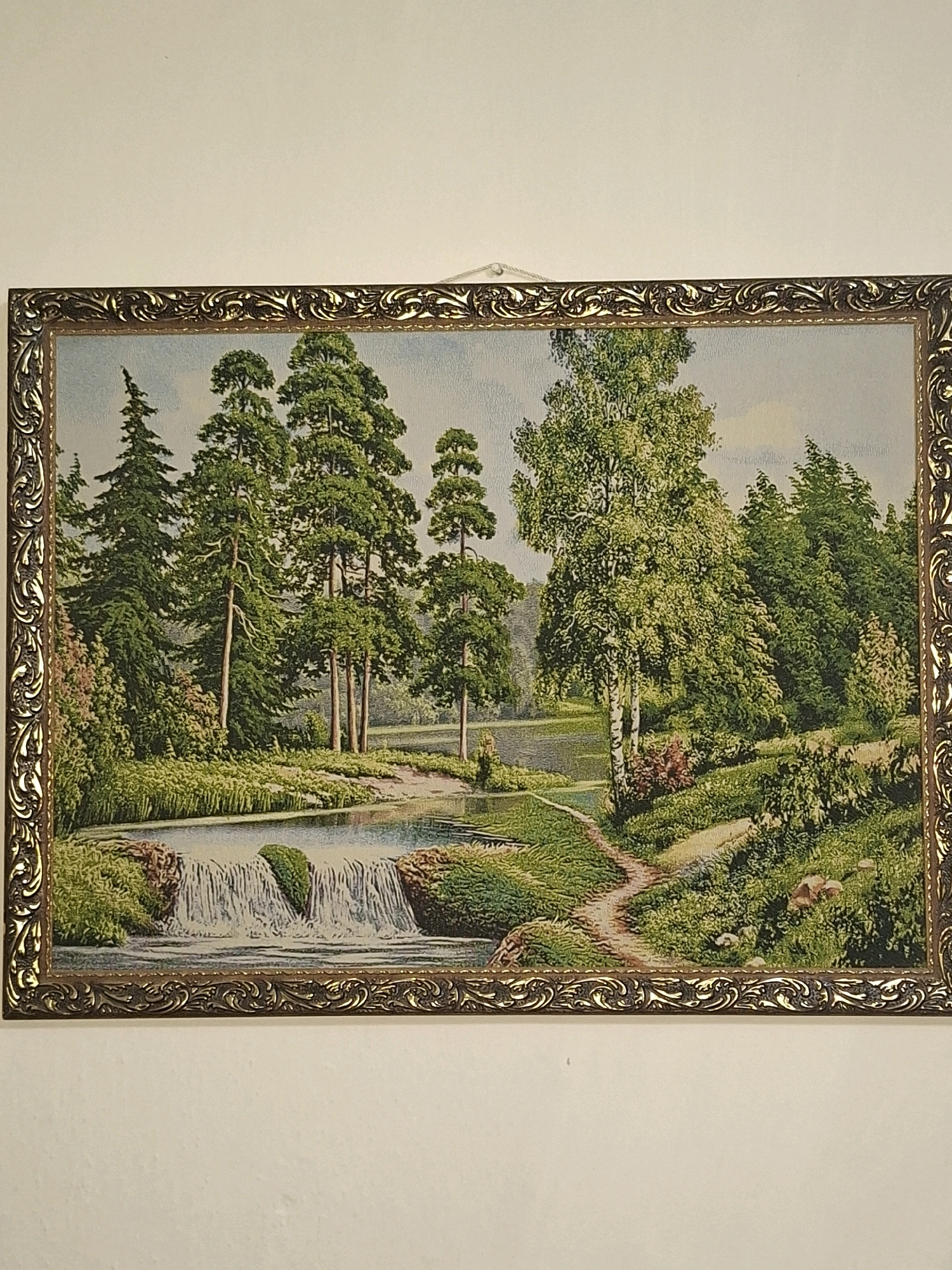 The image shows a framed tapestry depicting a serene landscape. A small waterfall cascades into a body of water in the foreground, bordered by lush green foliage and trees. A winding path leads through a wooded area with a variety of trees, including tall pines and a prominent birch tree with white bark. The scene evokes a sense of tranquility and natural beauty. The tapestry is hanging on a plain white wall. There is no discernible information or visual cue within the image that relates it to the city of Almaty, Kazakhstan.