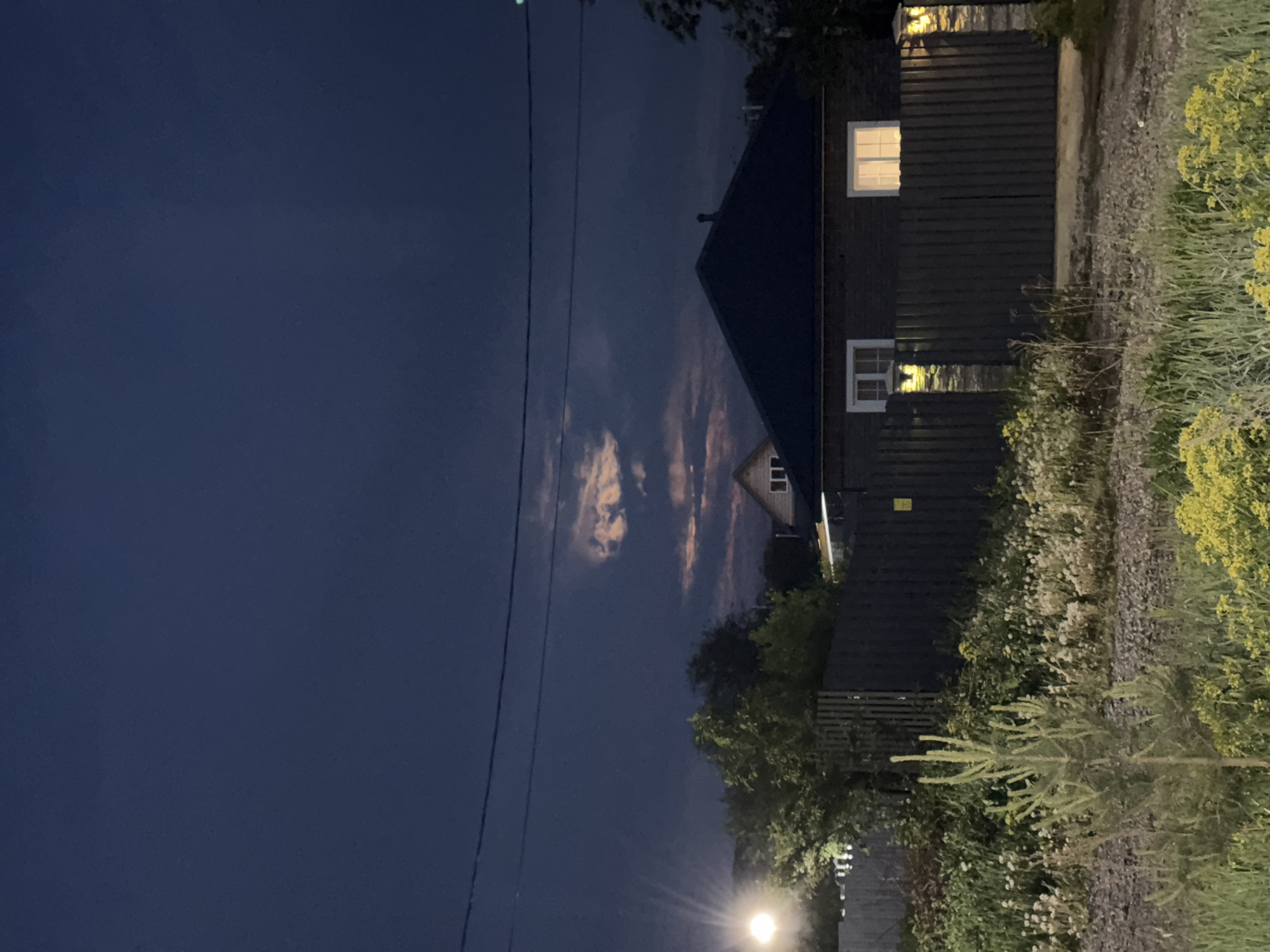 The image depicts a night scene in Горный, Russia. A wooden house is visible with lit windows, adjacent to a gravel path bordered by overgrown vegetation. In the foreground, power lines stretch across a dark blue sky with patches of clouds. A street lamp illuminates the scene from below.