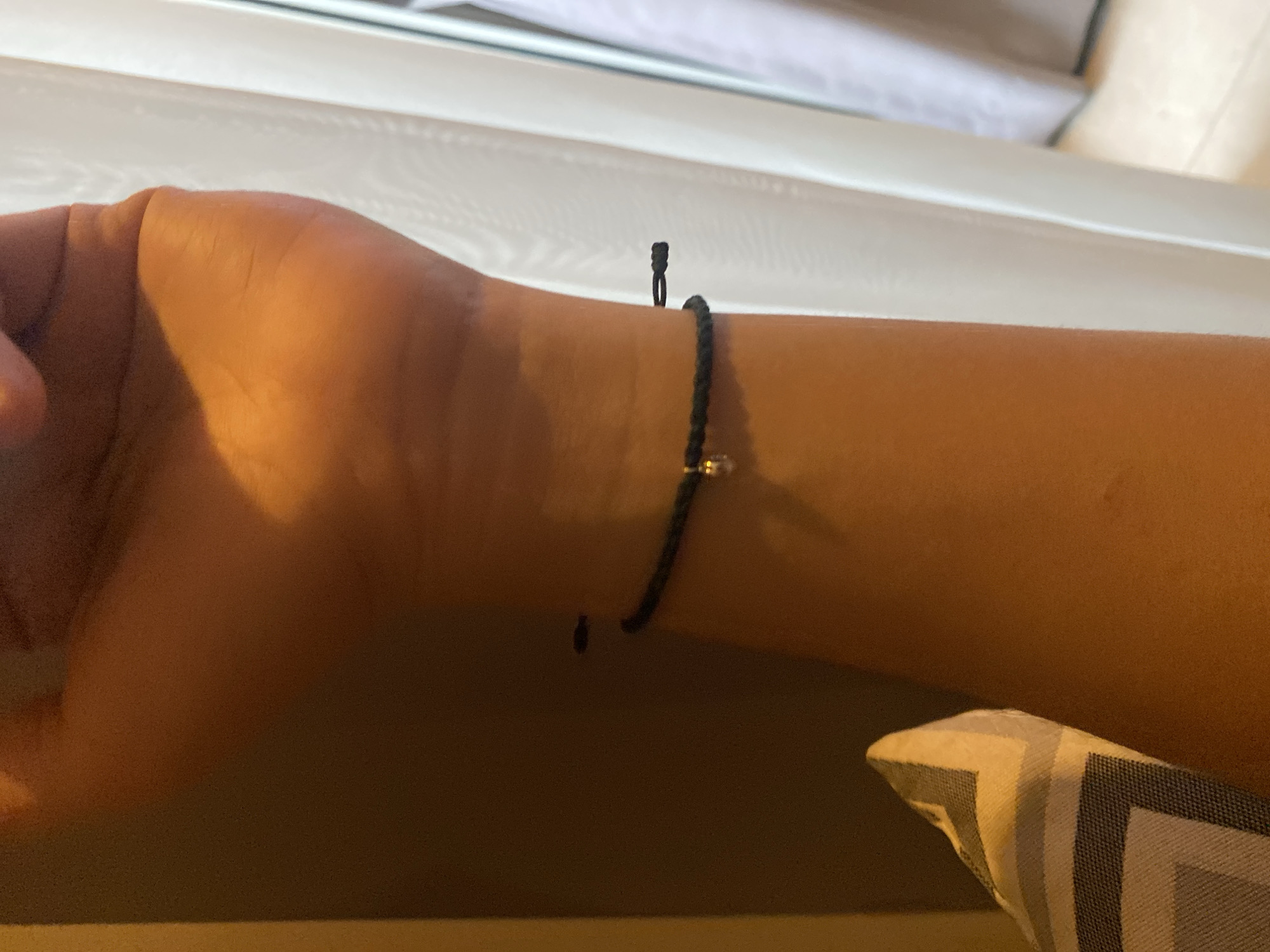 A close-up, low-angle shot shows a person's forearm and wrist wearing a thin, dark braided bracelet. The bracelet has a small, shiny charm dangling from it, possibly a bead or a small metallic object. The skin tone is warm and tanned. The background is dimly lit and out of focus, with a vertical pattern that appears to be a sheer curtain or blind on the right side of the frame, and a textured, patterned cushion on the left. The lighting suggests an indoor setting, likely during the daytime given the presence of natural light filtering through the curtain. There are no visible elements that definitively place this image within Central Jakarta, Indonesia.