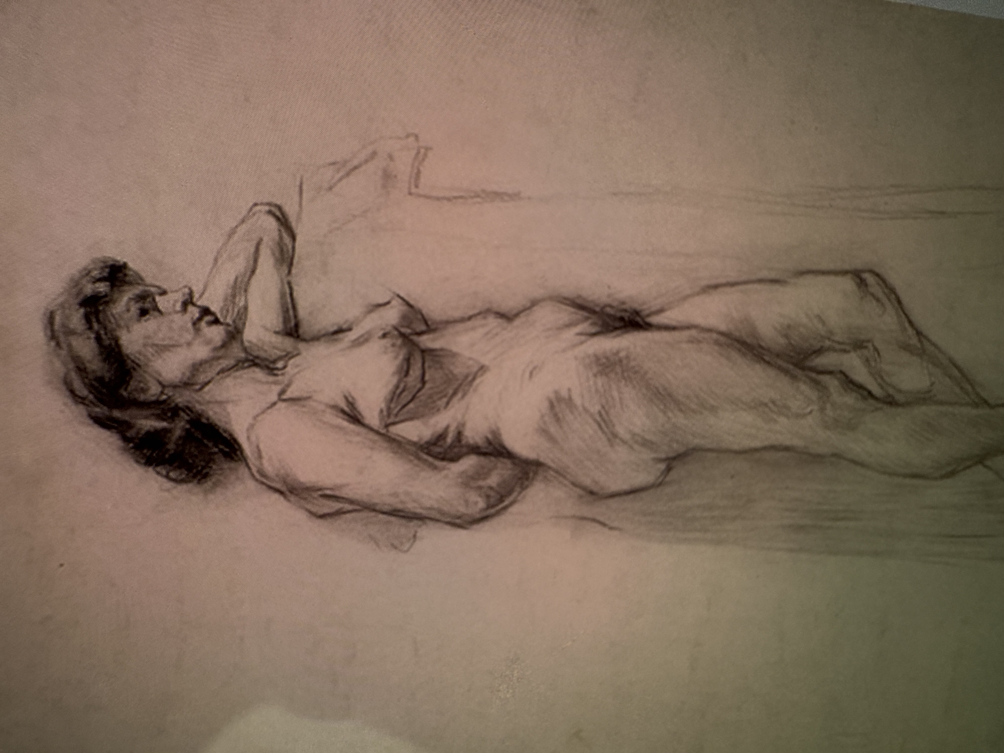 A charcoal drawing depicts a nude woman in a three-quarter view, leaning against a vertical, indistinct object. The woman's left arm is bent, supporting her head, while her right arm rests against the object. Her gaze is directed downwards and to her right. The drawing is rendered in monochrome with visible shading and texture. The background is plain and undetailed. The drawing is not located in a specific city or environment.