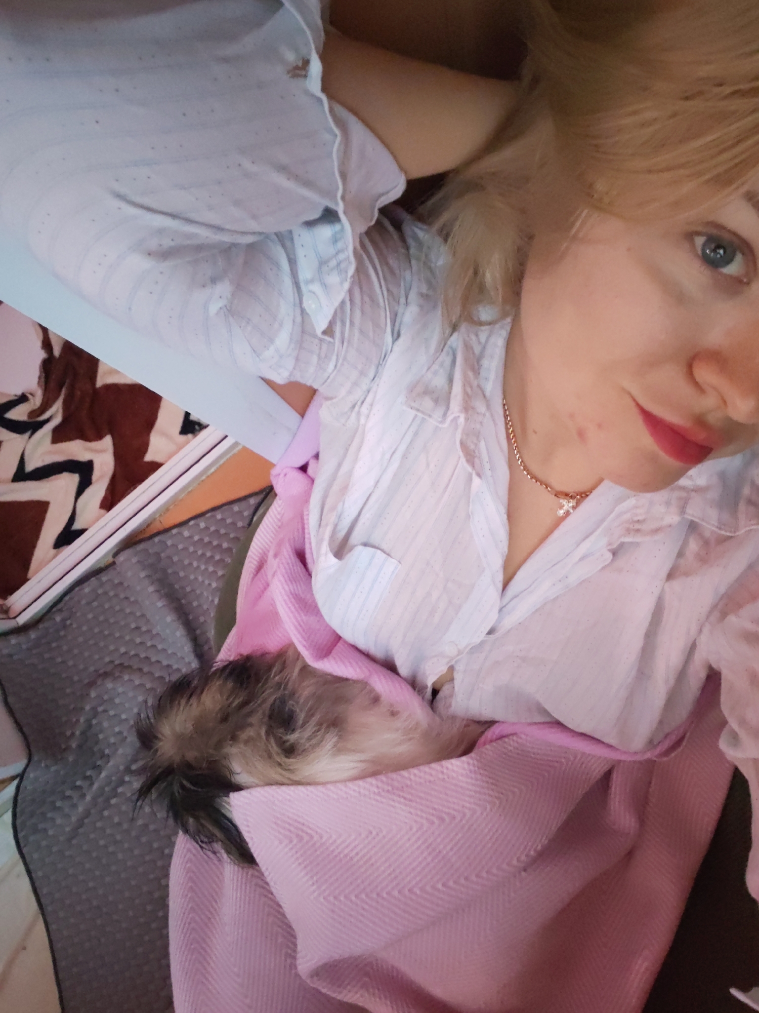 The image shows a close-up of a woman with blonde hair and blue eyes, wearing a white collared shirt. She is holding a small, fluffy dog, possibly a puppy, wrapped in a pink blanket. The woman is looking at the camera, and her face is partially visible. The dog's head is resting on the woman's chest, and only its fur is visible. The background shows a patterned blanket and a gray textured surface. The scene appears to be indoors. There is no identifiable information within the image to suggest a location in Izhevsk, Russia.
