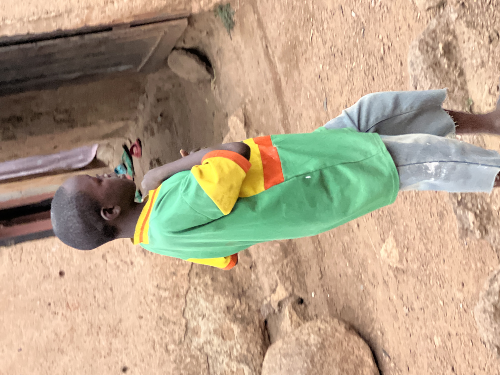 A young, dark-skinned child stands outdoors, viewed from the left side. The image is presented sideways, rotated 90 degrees clockwise from its natural orientation. The child wears a vibrant green t-shirt featuring prominent horizontal stripes of yellow and orange on the sleeves and across the upper chest. They are also dressed in light grey or faded blue trousers. The child's arms are slightly bent, and they appear to be looking upwards or towards their left with a neutral expression.
The setting consists of reddish-brown, sandy or earthy ground, which is uneven and textured. To the child's upper left (if the image were properly rotated), a section of a rough, light brown wall or structure is visible, featuring a dark, rectangular opening or recess. The bright, clear lighting indicates that it is daytime. The earthy tones and simple, robust construction of the background elements suggest a rural or semi-urban environment, fitting the context of Jos, Nigeria. No specific activity beyond standing is depicted, and no text is visible.