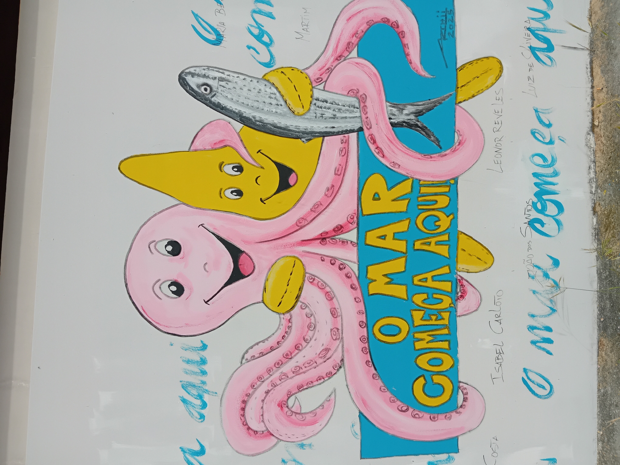 This is a vibrant and cheerful cartoon-style mural, likely painted on an outdoor wall or panel in Figueira da Foz, Portugal. The main subjects are a large, smiling pink octopus and a smaller, happy yellow star, both interacting with a realistic-looking silver fish.
The pink octopus, with its numerous tentacles, playfully holds the fish across its body using two of its limbs, which appear to have yellow cuff-like markings. Its round eyes and wide smile convey a jovial expression. Below the fish, the yellow star also sports a wide smile and large eyes, seemingly supporting or engaging with the fish. The fish itself is depicted with notable detail, including scales and a textured appearance, contrasting with the smooth, cartoonish style of the octopus and star.
A prominent vertical blue banner runs through the right side of the artwork, featuring bold yellow text that reads, "O MAR COMEÇA AQUI" (The Sea Starts Here), clearly indicating a marine theme and connection to the ocean. Around the central illustration, numerous signatures and names are written in blue and black, suggesting multiple contributors or dedications to the artwork. Visible names include "MARIA BE", "MARTIM", "LEONOR REVELES", "LUIZ DE OLIVEIRA", "JOÃO DOS SANTOS", "ISABEL CARLOTO", and "COSTA". A date, "2025", is also visible near the top right of the blue banner. The overall impression is one of a community art project, possibly for children or celebrating a local connection to the sea. The bright colors and playful expressions create a joyful and welcoming atmosphere.