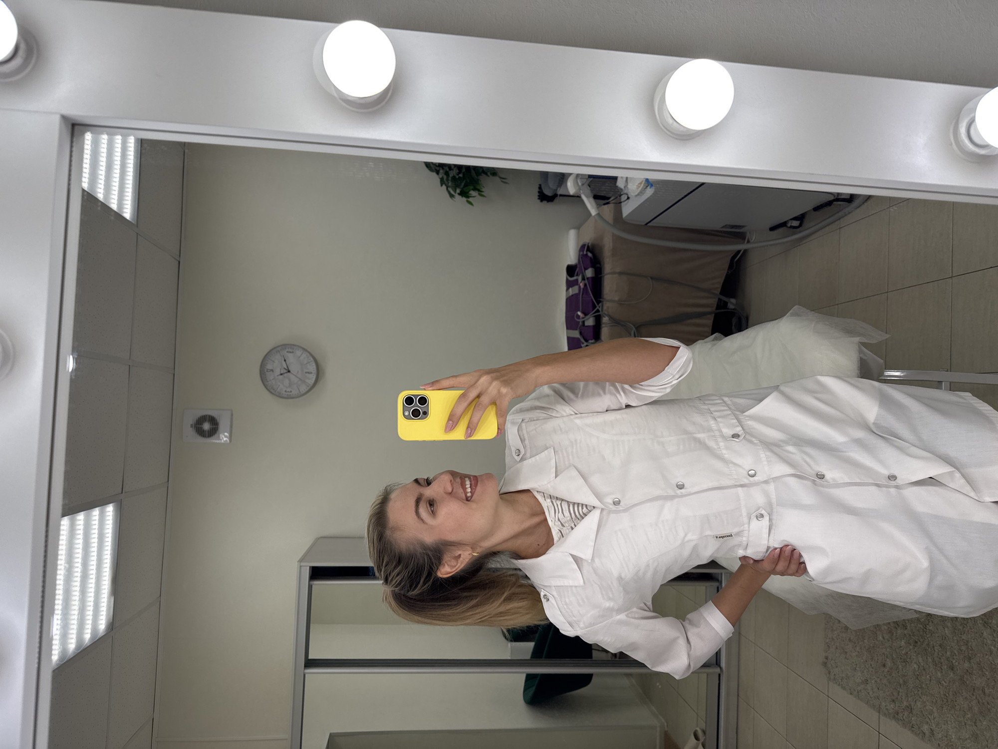 A woman in a white lab coat is taking a selfie in a mirror. The mirror is surrounded by round light bulbs. Behind the woman, a clock hangs on a pale wall. Medical equipment is visible in the background, along with a houseplant and white linens on what appears to be a medical bed. A sliding door closet is seen in the reflection of the mirror. The location is Unknown, Unknown.