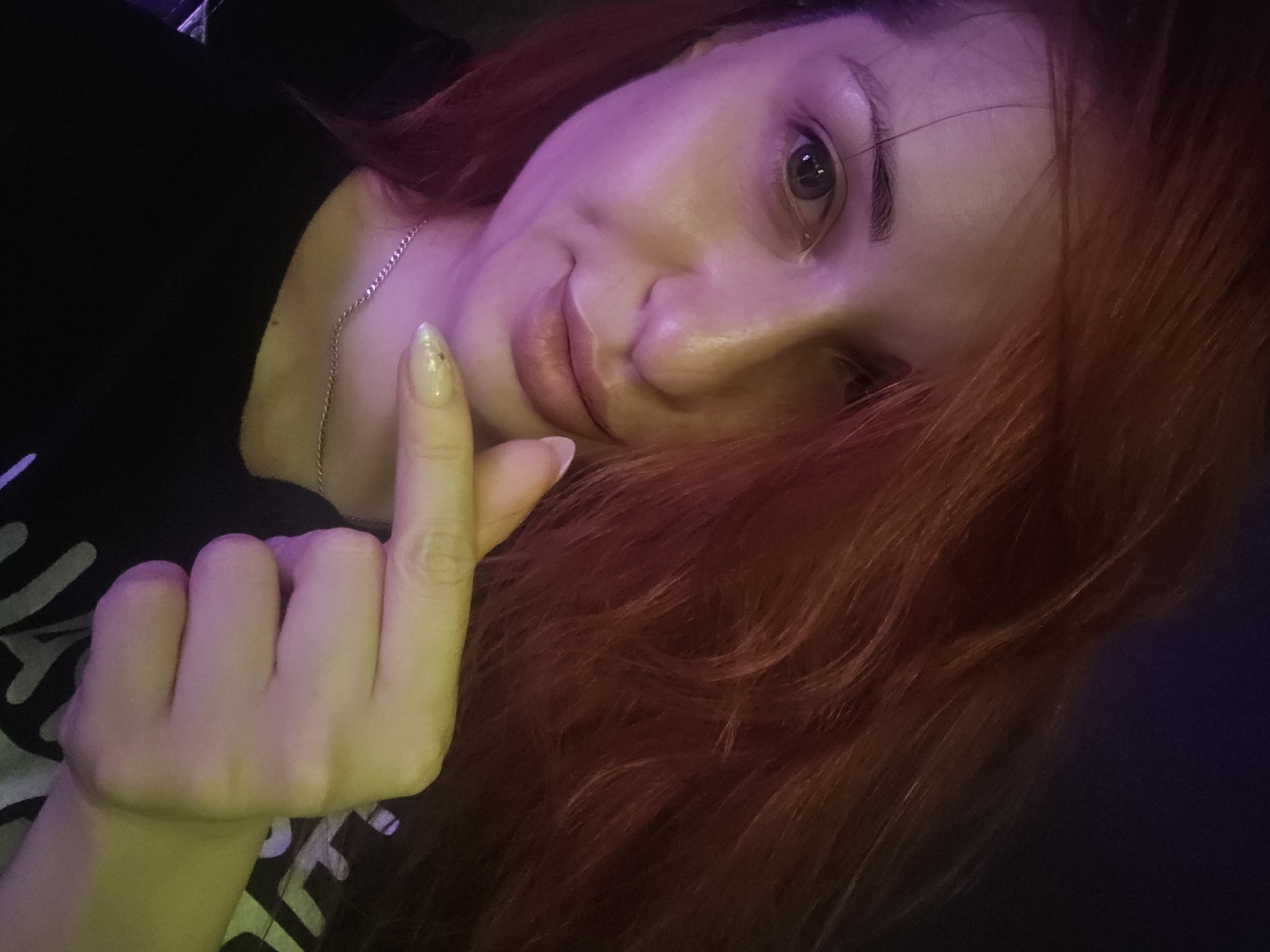 The image is a close-up selfie of a person with red hair. The person is wearing a black t-shirt and a silver necklace. Their right hand is raised, with the index finger pointing towards the camera. The background is dark and out of focus, with hints of purple and yellow lighting. The person's face is partially illuminated by this lighting. The image does not contain any identifiable landmarks or features that would indicate its location in Burgas, Bulgaria.