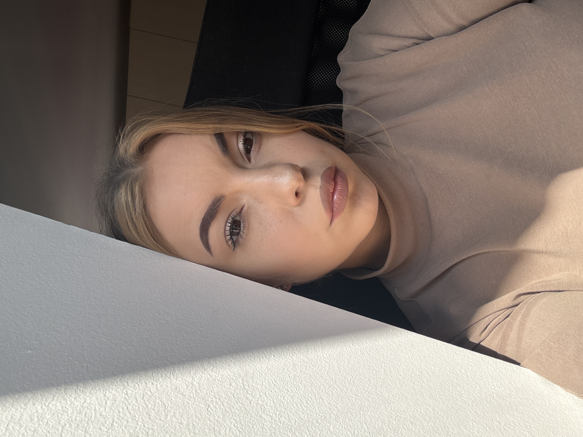 An indoor portrait depicts a young adult female subject, located in the city of Sofia, Bulgaria. The subject is positioned facing the camera, with her gaze directed forward. Her hair is blonde with visible darker roots, parted and falling to her right. She has dark brown eyes with visible mascara and light eyeshadow, and well-defined, dark eyebrows. Her lips are full and coated with a nude-toned lipstick. Her complexion is fair, with minor freckling visible on her nose and cheeks. A small earring is present in her left ear. She is wearing a light beige or brown long-sleeved top with a high neckline, made of a fine-knit or ribbed fabric. Strong natural light originates from the left side of the frame, illuminating the left side of her face and body and casting a distinct diagonal shadow line on the off-white, textured wall that occupies the left portion of the image. The background to her right is darker, revealing part of a black mesh office chair and dark brown architectural or furniture elements above it.