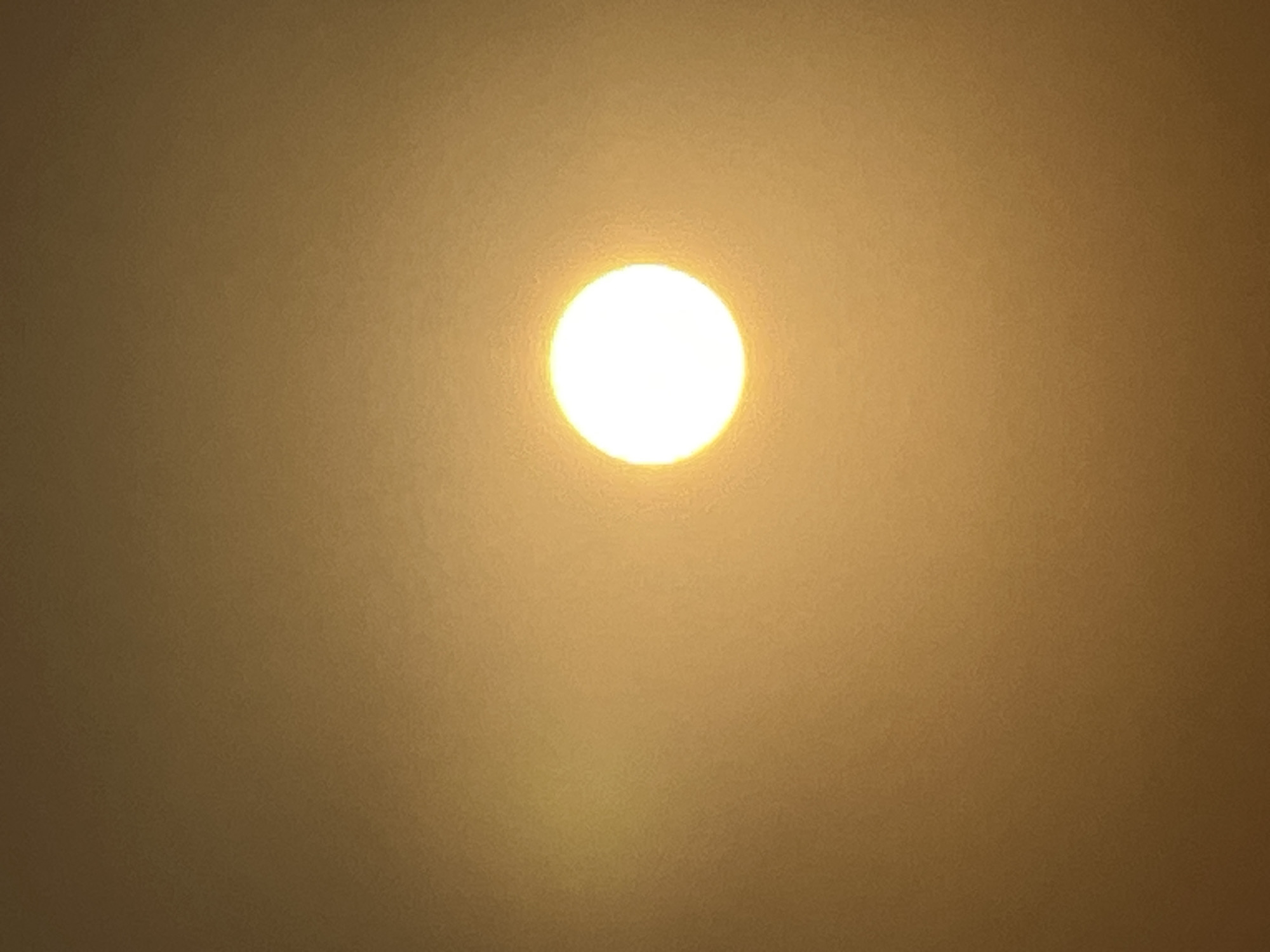 The image shows a bright, glowing sun partially obscured by atmospheric haze, set against a gradient of warm, muted tones. The sun appears as a distinct white-yellow circle in the upper right quadrant of the frame. The background is a soft, diffused wash of golden brown and orange, indicating a possibly hazy or dusty environment. There are no discernible landmarks or objects within the frame to provide specific context for a location in Kuta, Indonesia. The scene is devoid of any human presence or activity.