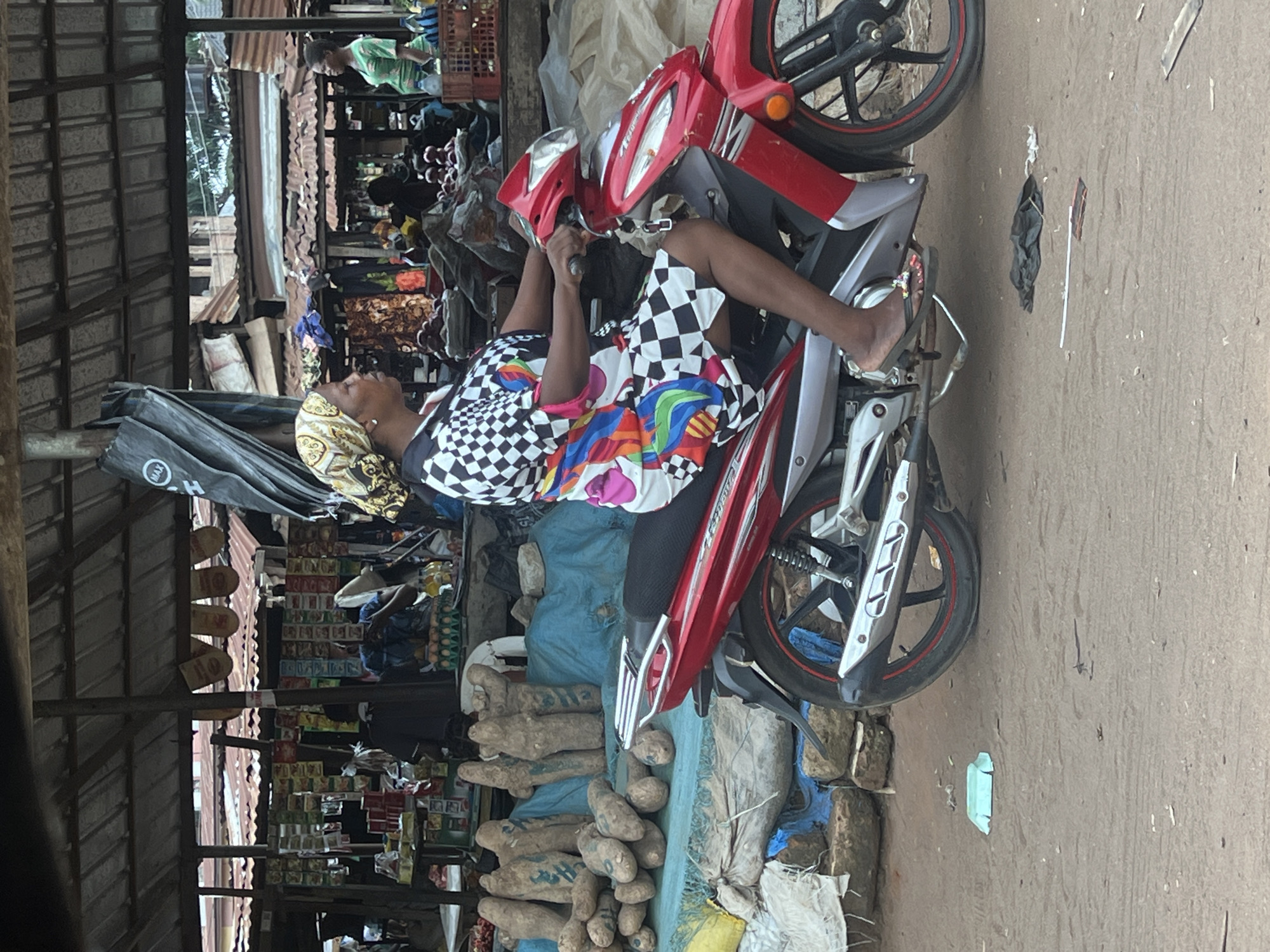 A market scene shows a woman seated on a red and silver motorcycle, surrounded by various items for sale. The woman wears a colorful patterned dress and a headscarf. The motorcycle is parked near piles of what appear to be yams. A store with shelves stocked with goods is visible in the background. The location is a market in an undefined city, Unknown.
