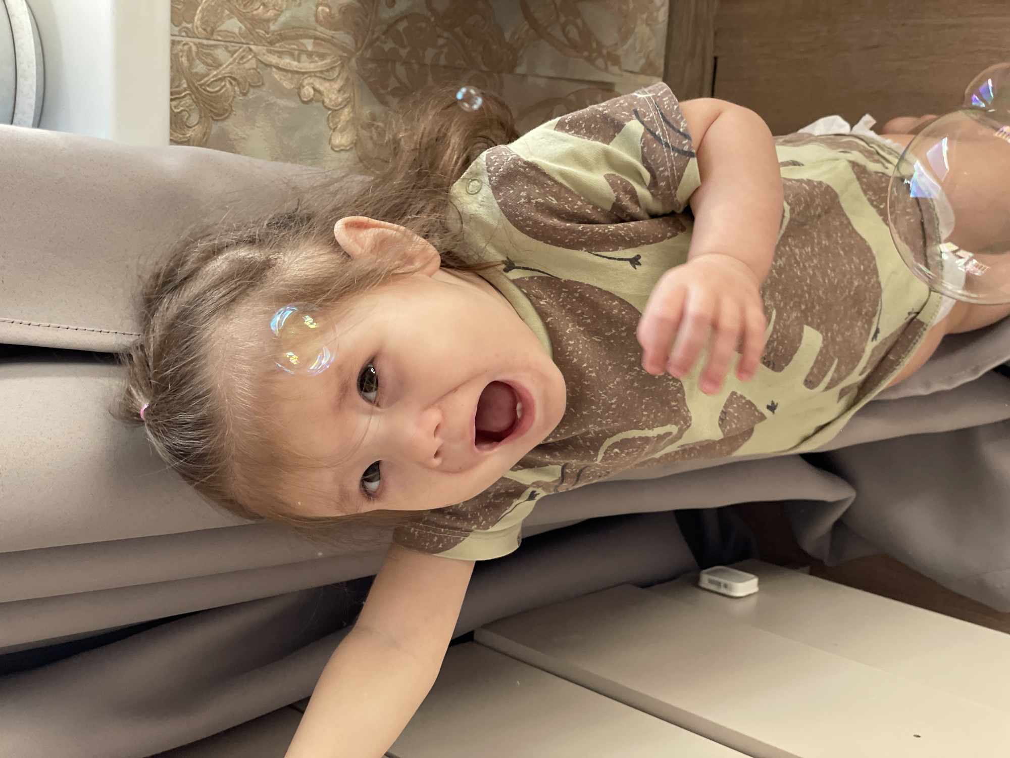 A young child lies on a light gray upholstered surface with a matching light gray panel behind it. The child, who is angled towards the camera, has light brown hair with a pink hair clip and is wearing a light yellow-green and brown patterned shirt. Their mouth is open as if shouting, and soap bubbles are visible near their face. A white object is on a white horizontal surface below the upholstered surface. More soap bubbles are visible in the upper right of the image. The location is unknown.