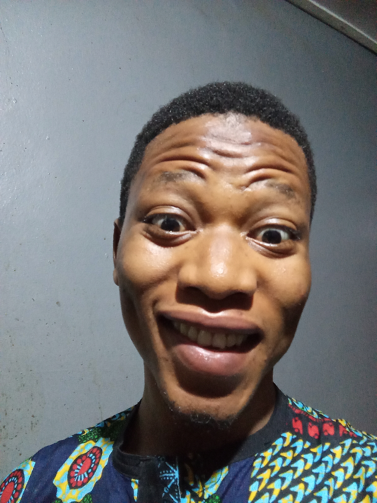 A head-and-shoulders shot shows a young man with dark skin and short, dark hair against a gray wall. He is smiling widely, showing his teeth, and his eyes are wide. He is wearing a colorful shirt with a blue background and patterns featuring red, yellow, and green elements. No elements suggesting a location in Oshogbo, Nigeria, are visible.