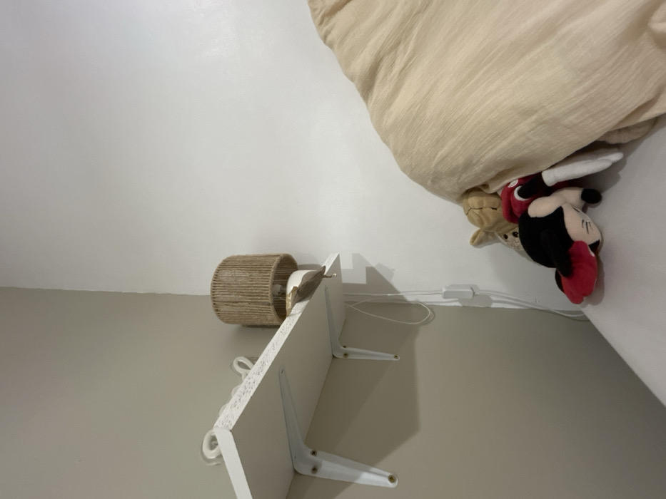 The image shows a corner of a room with a bed, a bedside lamp, and a white shelf. The bed has light beige bedding. Under the edge of the bedding are two plush toys, a brown one and a Mickey Mouse toy. The bedside lamp has a woven cylindrical shade. The white shelf is mounted on the wall with white brackets. A white cable runs along the wall near the shelf and the bed. The walls are painted in light grey and white.