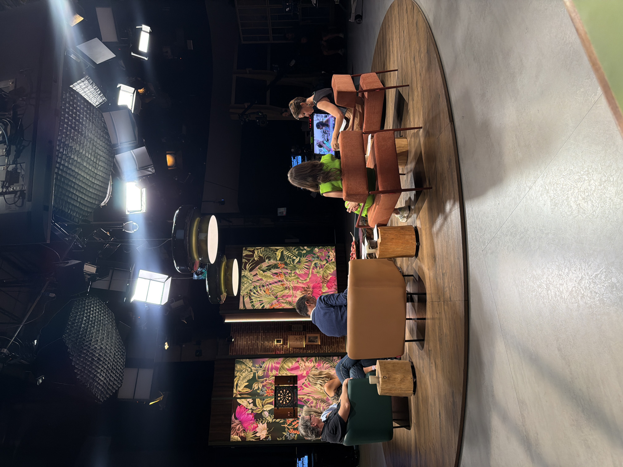 The image depicts a television studio set, likely located in the city of Sant Joan Despí, Spain, known for its media production facilities. The set features a circular wooden platform on a concrete floor, with several seating arrangements. In the background, two large panels display vibrant tropical foliage with pink and green hues. A dartboard is mounted on the left panel.
Five individuals are present:
* On the left, a man with graying hair is seated in a dark green armchair, facing inward. Next to him, a woman with blonde hair is also seated in the armchair, her body angled towards the center.
* In the middle, a man in a blue suit is seated in a tan armchair, facing away from the camera.
* On the right, two women are seated in tan armchairs. One woman, facing away from the camera, wears a bright green outfit. The other woman, facing towards a monitor, wears a black sleeveless top and a light brown skirt.
Several objects are visible:
* Two cylindrical wooden stools are placed near the armchairs.
* A low-profile coffee table with some items on it is situated in the center of the seating arrangement.
* A large monitor is positioned on a stand behind the two women on the right.
* Studio lighting equipment, including large softboxes and overhead lights, is visible in the dark ceiling.
* Camera equipment and other production elements are present in the background, suggesting an active filming environment.
The overall scene suggests a talk show or panel discussion being recorded in a professional studio setting.