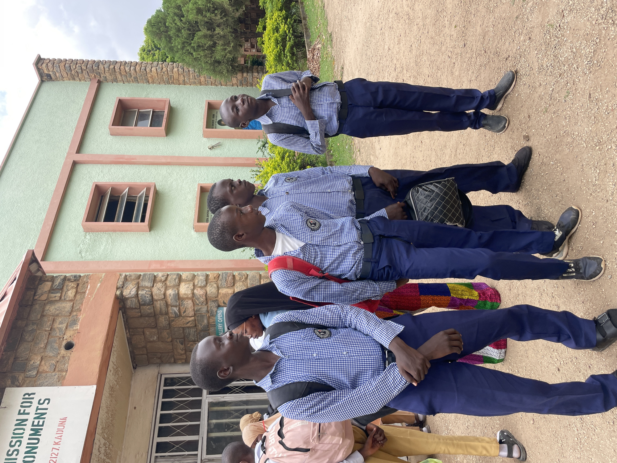 The image depicts a group of young people, likely students, standing outside a light green building with a stone facade. The building has several windows and appears to be a public institution, possibly a museum or gallery, as indicated by a sign that reads "ISSION FOR MONUMENTS" and "2127 KADUNA".
The subjects are all dressed in uniforms consisting of blue and white checkered shirts and dark blue trousers. Most are wearing backpacks. Some of the individuals are looking up and to the side, suggesting they are waiting or observing something. The overall mood seems neutral and organized.
The scene is set outdoors on a dirt ground with scattered gravel, under what appears to be a cloudy sky. There are trees and bushes in the background, indicating a location with some greenery. The lighting suggests it is daytime. The orientation of the image is on its side.