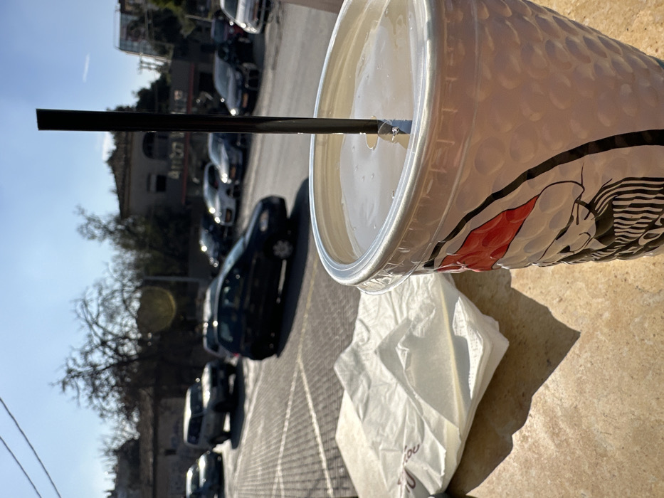 An outdoor, slightly tilted, low-angle shot captures a large, textured white disposable cup with a black straw in the foreground, filled with a light-colored liquid. The cup rests on a light-colored paved or tiled surface, casting a prominent shadow, indicating bright sunlight and clear skies during the day. A crumpled white plastic bag is visible next to the cup, with faint Greek text "σου" (you) printed on it, suggesting a location in Greece.
The cup features a distinctive dimpled texture across its surface and a graphic design with red and black elements on its side, partially visible, appearing to depict a stylized figure or logo. In the out-of-focus background, a street or parking lot is visible, lined with several parked cars. Further back, buildings and bare trees are present under a clear blue sky. The scene depicts a casual outdoor setting, possibly a street-side cafe or takeaway spot in Metamorfosi, Greece.