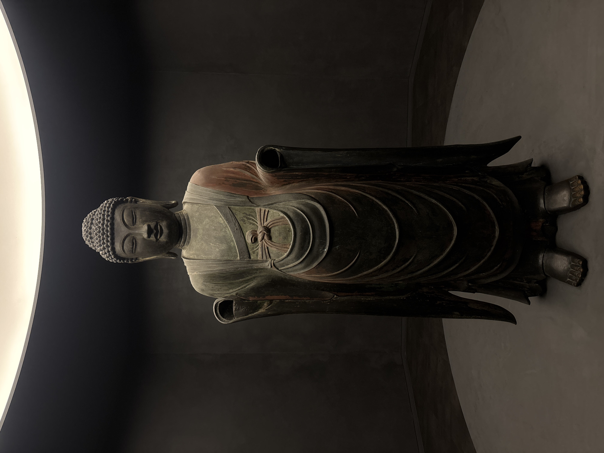 A full-body, front-facing bronze statue of Buddha stands in a dimly lit, circular room. The statue is positioned on a circular platform, with a subtle spotlight illuminating it from above. The background consists of dark, textured walls that curve away from the statue. The Buddha statue is depicted in a standing pose, with its right hand raised and palm facing outward, and its left hand lowered. It wears traditional monastic robes, with one shoulder exposed and draped with a reddish-brown fabric. The statue has a serene expression with closed eyes and a coiled ushnisha on its head. The lighting casts long shadows, emphasizing the statue's form and the texture of the robes. The overall atmosphere is solemn and contemplative. This statue is located at Bulguksa Temple in Gyeongju-si, South Korea.