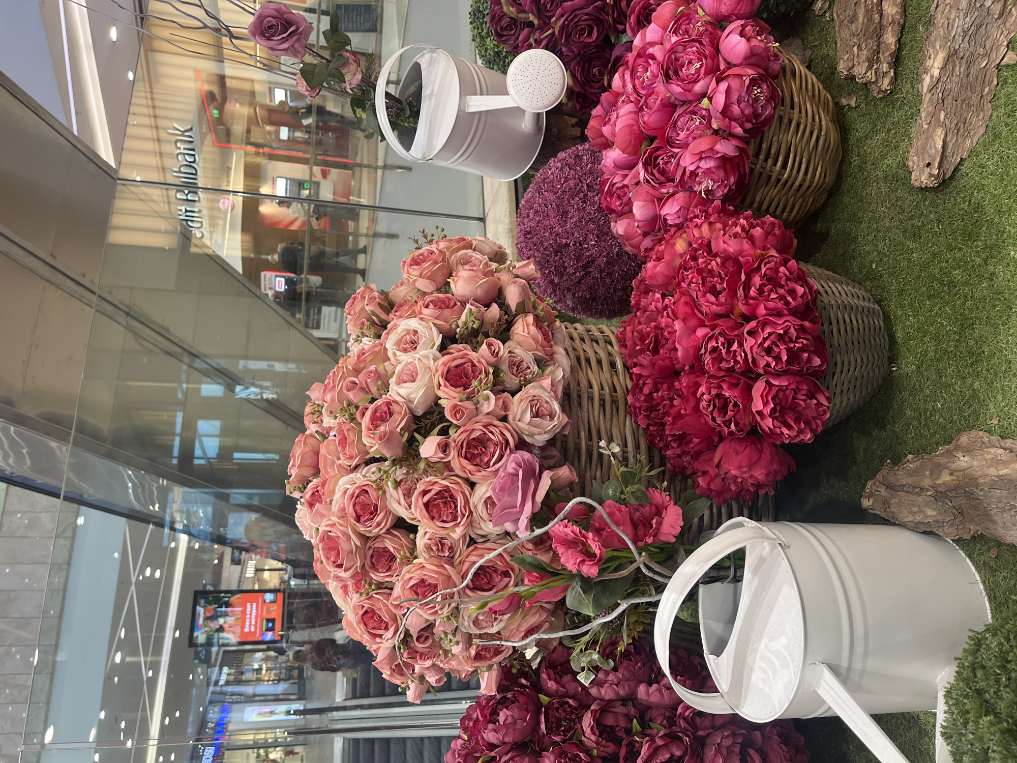 The image depicts a floral display in a commercial setting, possibly a shopping mall, in Sofia, Bulgaria. The foreground features arrangements of pink and red artificial flowers in woven baskets, interspersed with white watering cans. The flowers consist of roses, peonies, and a purple pompon-like flower. The display sits on a patch of artificial green grass, accented with pieces of bark.
The background reveals a glass facade reflecting the interior of the mall. Reflected signs indicate the presence of "Credit Bulbank," a bank, and digital displays are visible. Blurred figures of shoppers and mall employees are visible in the reflections. The lighting is bright and artificial, typical of an indoor commercial space.