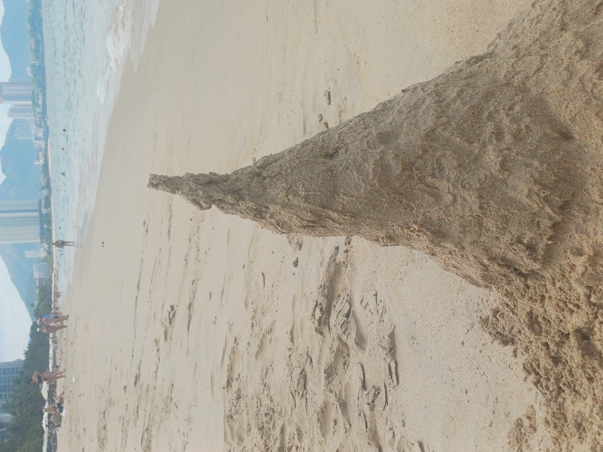 The image depicts a beach in Nha Trang, Vietnam. In the foreground, a sandcastle stands prominently, its conical shape sculpted from damp sand. The sandcastle's surface displays a rough, textured appearance, indicating it was recently constructed. The beach is composed of fine, light-colored sand with subtle undulations and ripples. In the middle ground, several people are visible, some walking along the shoreline and others relaxing on the beach. The water of the ocean is a muted blue-green, with gentle waves lapping at the shore. In the background, the cityscape of Nha Trang rises against a hazy sky, characterized by modern buildings and distant mountains. Palm trees are scattered along the coastline, adding a tropical element to the scene. The overall atmosphere suggests a warm, sunny day at a popular tourist destination.