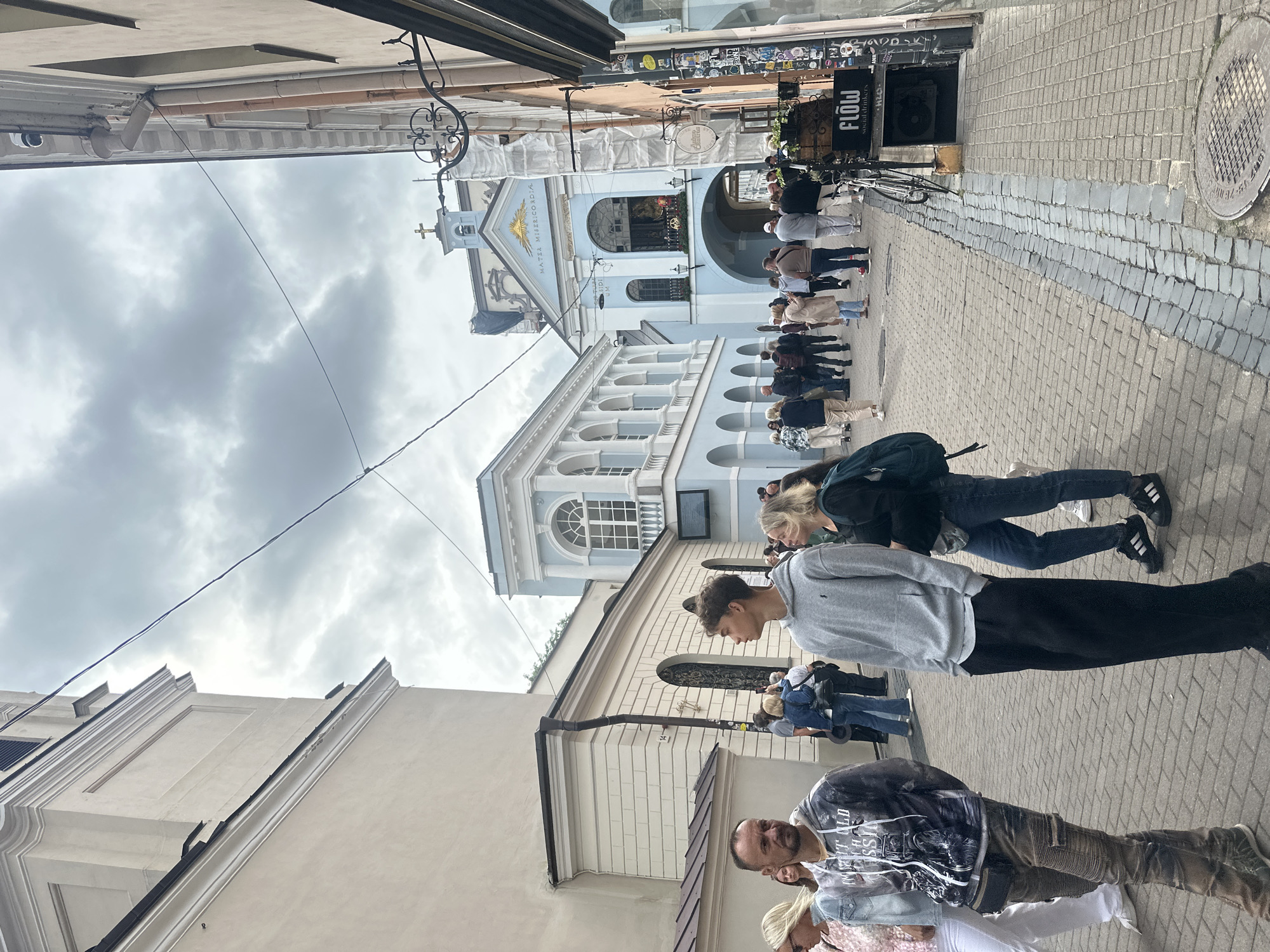 The image depicts a street scene in Vilnius, Lithuania, likely in the Old Town, characterized by cobblestone pavement and historic architecture. In the foreground, several individuals are walking down the street. Two young men are prominently positioned, one in a grey hoodie and black pants, facing right, and another in a graphic hoodie and patterned pants, facing left. A group of people, including women and men, are seen further down the street, some walking, others standing and observing.
Dominating the background is a light blue building with arched windows and colonnades, a common architectural style in European cities. Above this structure, a smaller, more ornate blue building with a cross on top, likely a chapel or part of a larger religious complex, is visible. The sky is overcast with clouds, suggesting a temperate climate. Electrical wires are strung across the upper portion of the frame, typical of urban environments. On the right side of the street, a shopfront with signage is present, including a black sign that reads "FLOW social drinkers." A bicycle is propped against a wall near the shop. The overall atmosphere is one of a bustling urban thoroughfare with a blend of historical and contemporary elements.