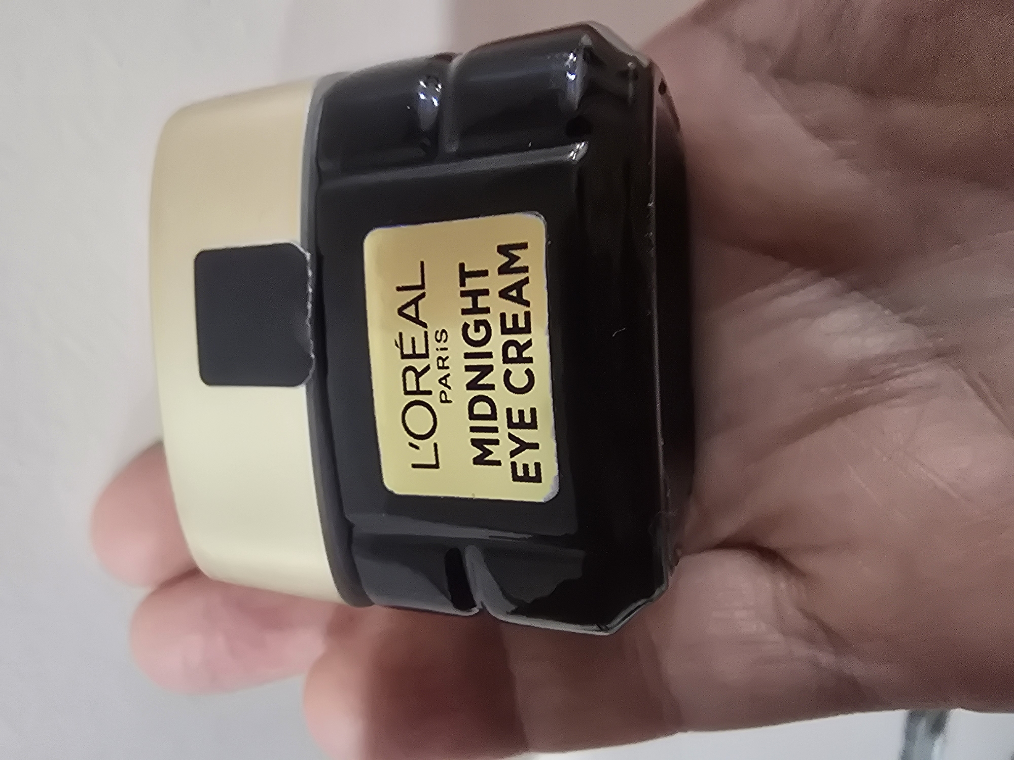 A black jar of L'Oréal Midnight Eye Cream is held in a person's hand. The jar has a gold lid and a yellow label with black text that reads "L'ORÉAL PARIS MIDNIGHT EYE CREAM". The background is blurred and appears to be a white surface. The image does not contain information that would allow for its location to be determined in Ulan-Ude, Russia.