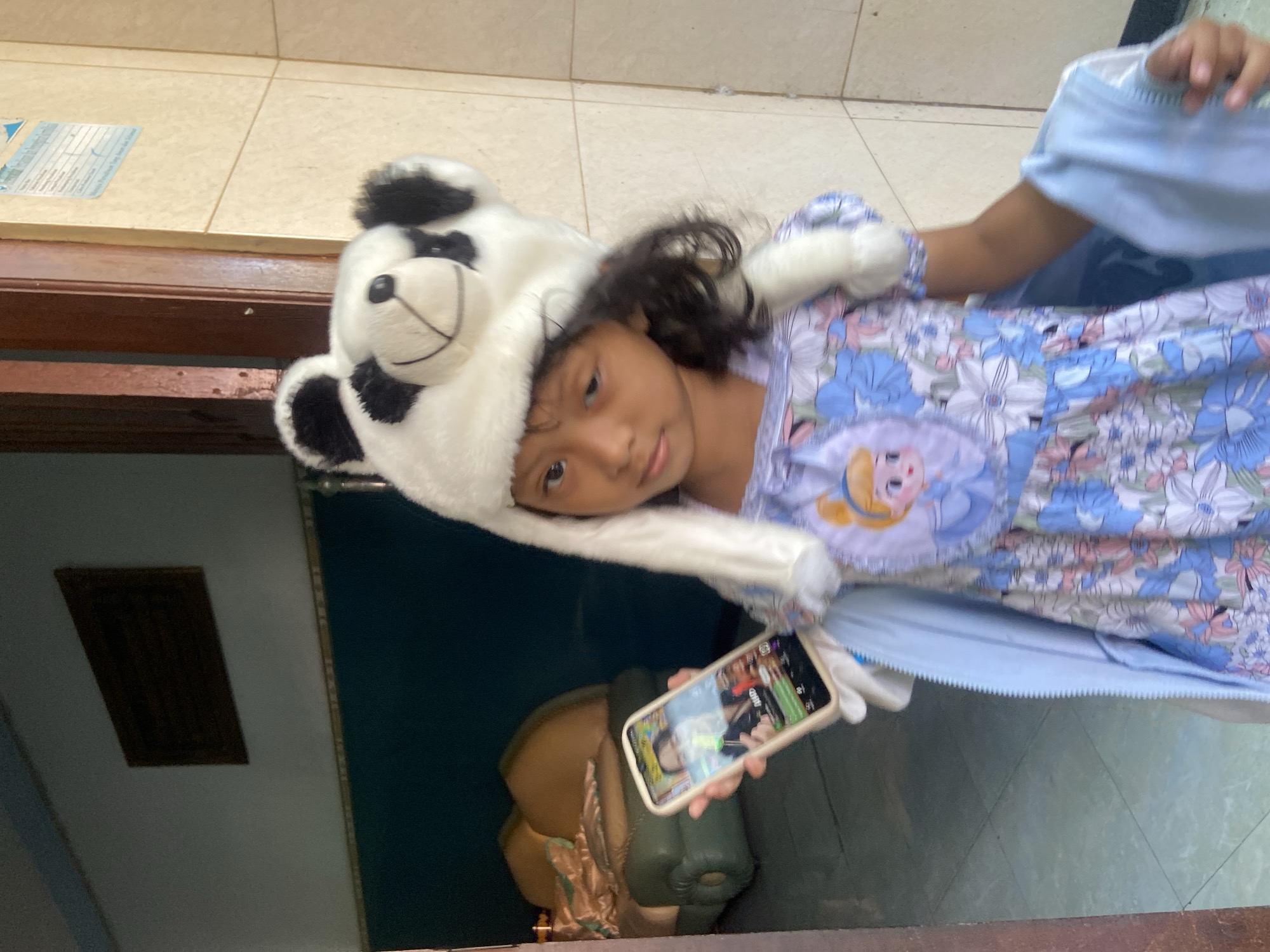 A young girl wearing a panda hat and a blue floral dress stands in what appears to be the entrance of a building. She is holding a smartphone in her left hand, displaying a video call. The background suggests an indoor setting with a doorway, tiled walls, and a couch visible in a darker room behind her. The location is identified as Gelam, Indonesia.