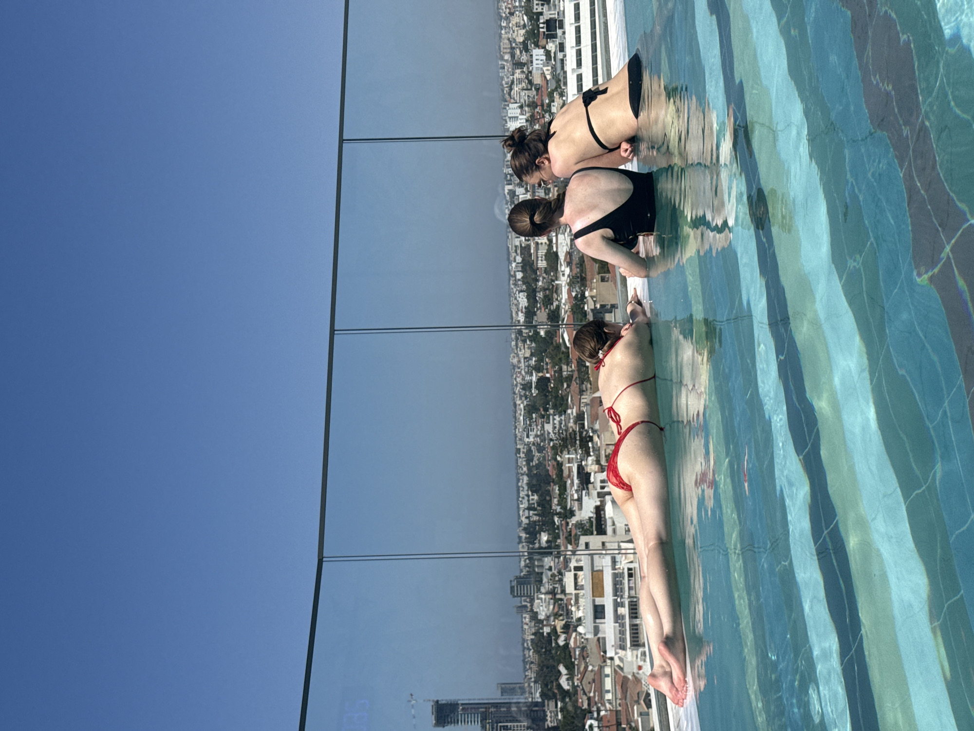 Three women are in a rooftop swimming pool overlooking the city of Limassol, Cyprus. The woman in the foreground is lying on her stomach in the water, wearing a red bikini. The two women behind her are leaning on the edge of the pool, facing away from the camera. The woman in the middle is wearing a black one-piece swimsuit, and the woman on the right is wearing a black bikini. The pool has a tiled bottom with stripes of blue and green. The city skyline is visible in the background, with buildings and trees. The sky is clear and blue.