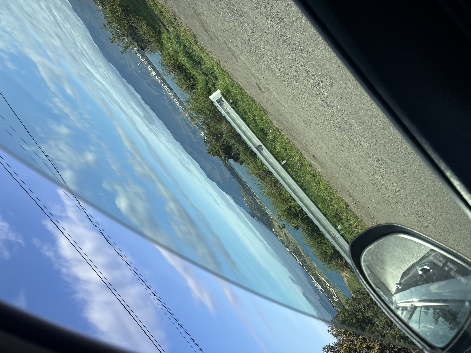 The photograph, taken from inside a vehicle, shows a landscape with a river running alongside a road, likely in a mountainous area. The sky is blue with scattered clouds, reflected in the car's window. Power lines are visible in the sky. A guardrail lines the road. A side-view mirror of the car is visible in the foreground. The specific location in Tbilisi, Georgia cannot be determined from the given image.