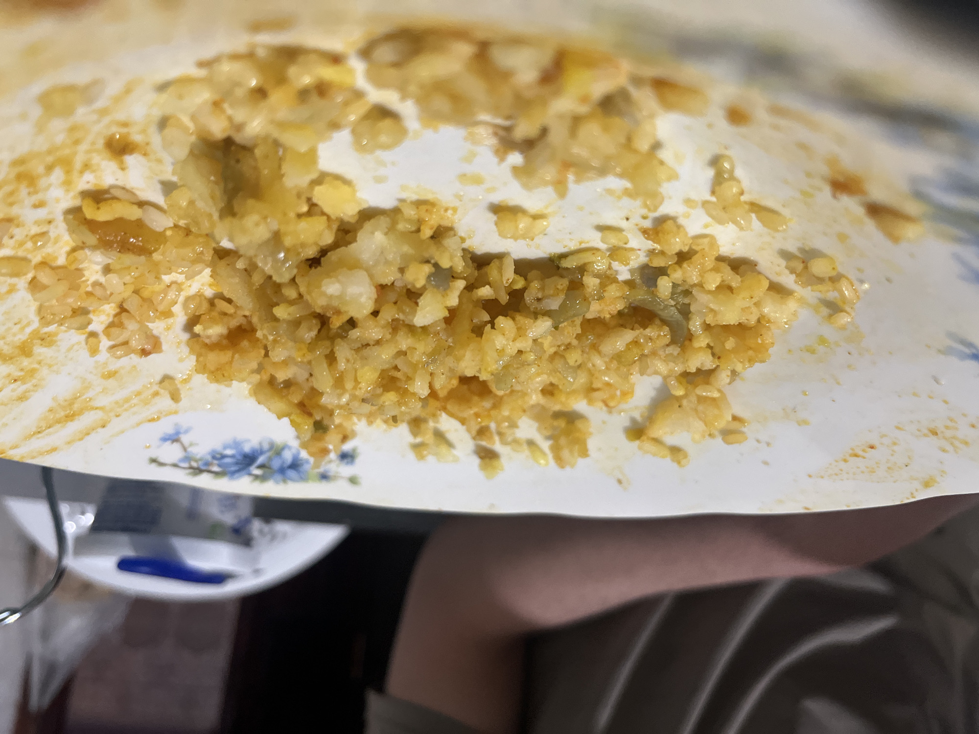 The image shows a partially eaten plate of rice, with a yellow/orange hue, possibly due to spices or sauce. The plate is white with a blue floral design. A small portion of a leg and part of clothing are visible in the lower right corner, suggesting the person is seated. There is a fork and a small package (possibly sauce) on a plate near the bottom left. The setting seems to be indoors. There are no indicators pointing to Ouaouizert, Morocco.