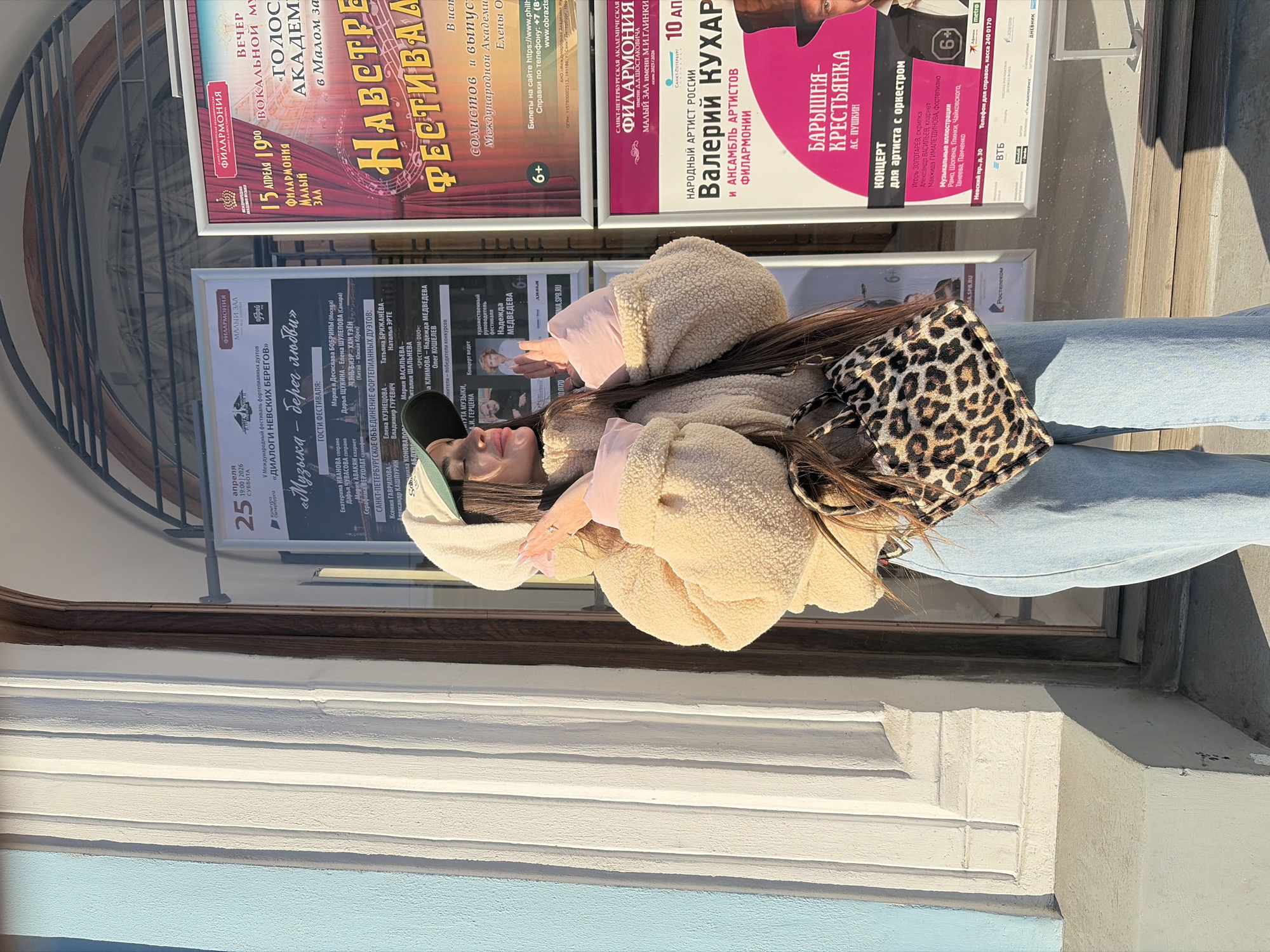 A woman is standing in front of a series of concert posters in Saint Petersburg, Russia. The posters advertise upcoming performances at the Saint Petersburg Academic Philharmonia. Specific performances include a vocal music evening on April 15th, a concert featuring Valery Kukhar, and a performance of "The Peasant Lady" by Alexander Pushkin. There is also a festival of piano duets titled "Dialogues of the Neva Banks". The woman is wearing a beige sherpa jacket, light blue jeans, and a cap, and carrying a leopard print bag. The building is of classical architecture, with a prominent facade and window.