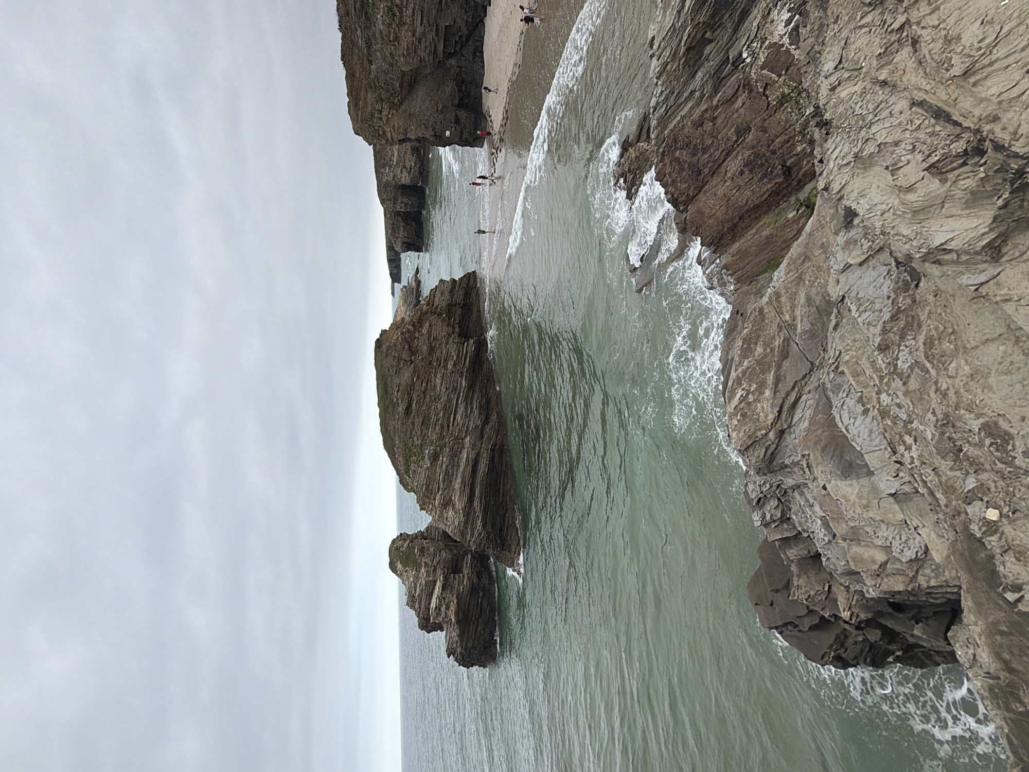This image captures a scenic view of Praia das Catedrais (Cathedral Beach) in Ribadeo, Spain, under an overcast sky. The beach is characterized by its dramatic rock formations, sculpted by the sea into arches and caves that resemble a cathedral. In the foreground, rough, stratified rock formations descend towards the water. The sea is a muted greenish-blue, with white waves crashing against the rocks and receding on the sandy shore.
Several people are visible on the beach and in the shallow water. Some are walking along the sand, while others are wading near the water's edge. A few figures are also seen near the base of the larger rock formations. The overall impression is one of a natural, rugged coastline with human activity present but not dominant. The lighting is diffused, typical of a cloudy day, which softens the colors and highlights the textures of the rocks and water.