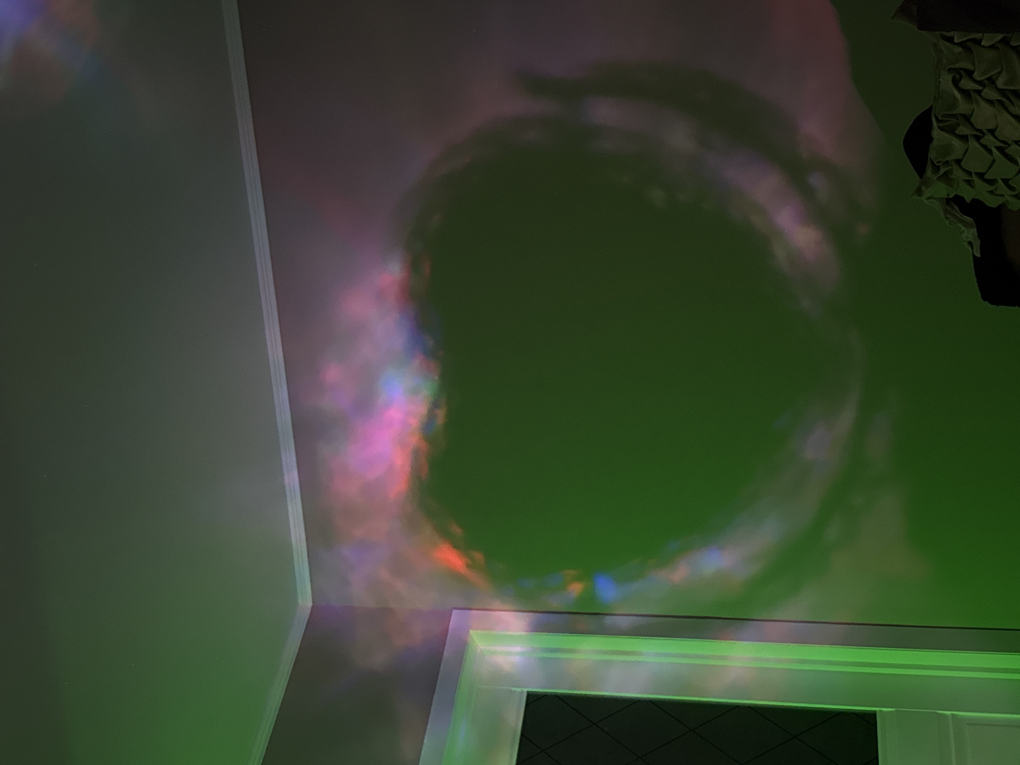 The image shows a room interior with a section of a wall, ceiling, and doorway visible. The wall is painted a light green, and the ceiling features a swirling, nebula-like projection with green, pink, red, and blue hues. The doorway, which appears to be at the bottom of the frame, has a white frame with green lighting accents. The visible interior of the doorway reveals a dark surface with a geometric pattern. A dark object or shadow is partially visible in the upper right corner. The location is unknown.