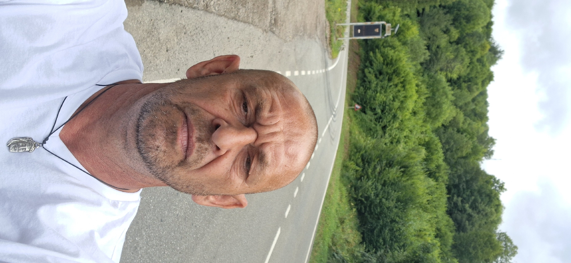 A selfie of a bald, middle-aged Caucasian man with a goatee and a furrowed brow is taken from the chest up, in front of a winding road. He is wearing a white t-shirt and a pendant necklace with a depiction of Jesus Christ. The road is paved with white dashed lines and a solid white line. To the left of the road, there is a tall signpost with a solar panel on top, and further down the road, a triangular warning sign. The background consists of a dense forest of green trees under a cloudy sky. The location is a rural area, possibly a mountain road, in an unknown city.