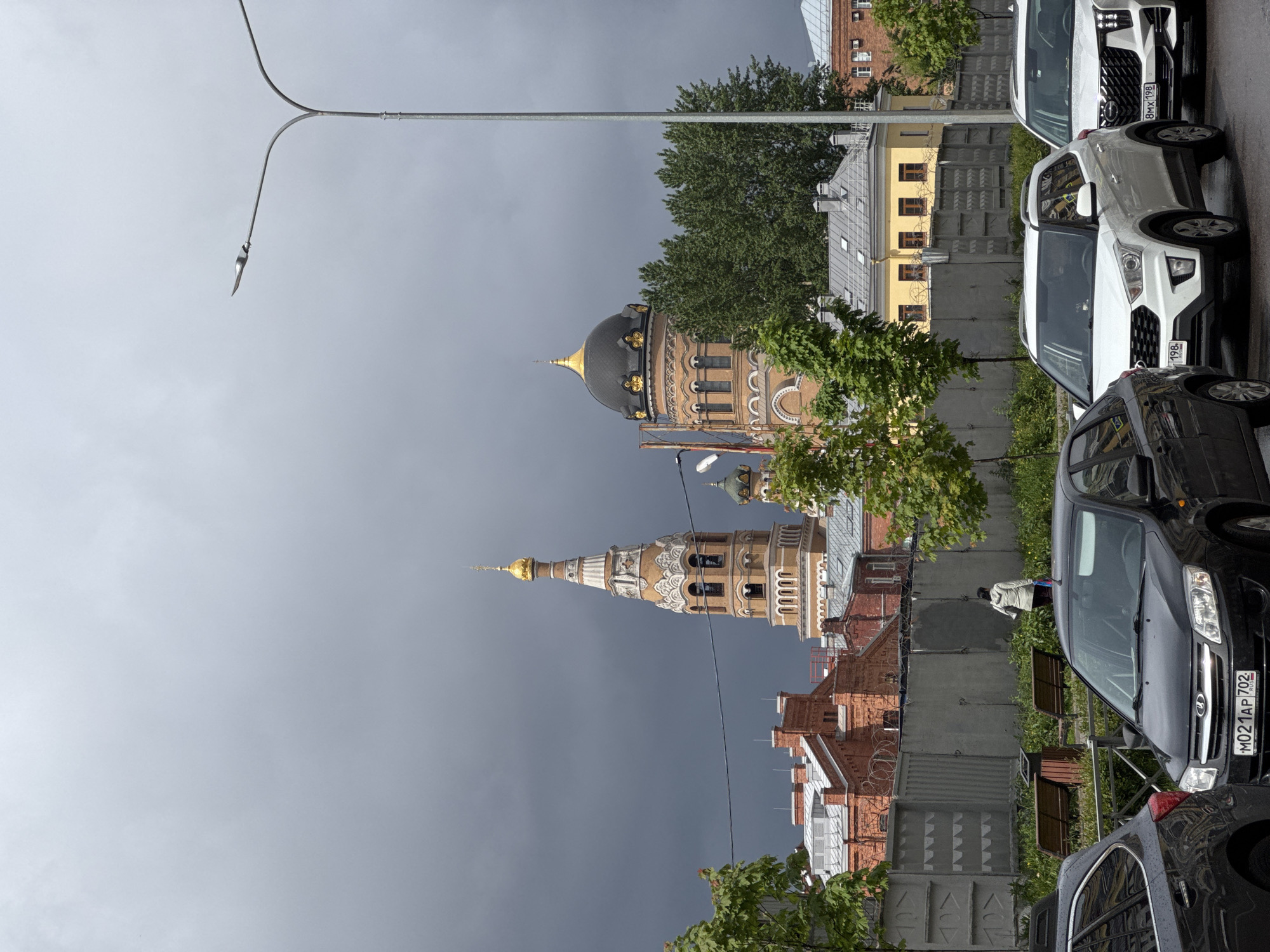 The image shows a view of a cityscape in Saint Petersburg, Russia. A tall, ornate church tower with a golden spire and a larger building with a golden dome are prominent features. The sky is overcast and gray. In the foreground, a row of parked cars lines the street. A streetlight pole is visible on the left. The buildings and rooftops are varied in style and color, with some having brick facades. A concrete wall or fence separates the street from the buildings. The scene appears to be taken on a street near a historical or religious landmark in the city.