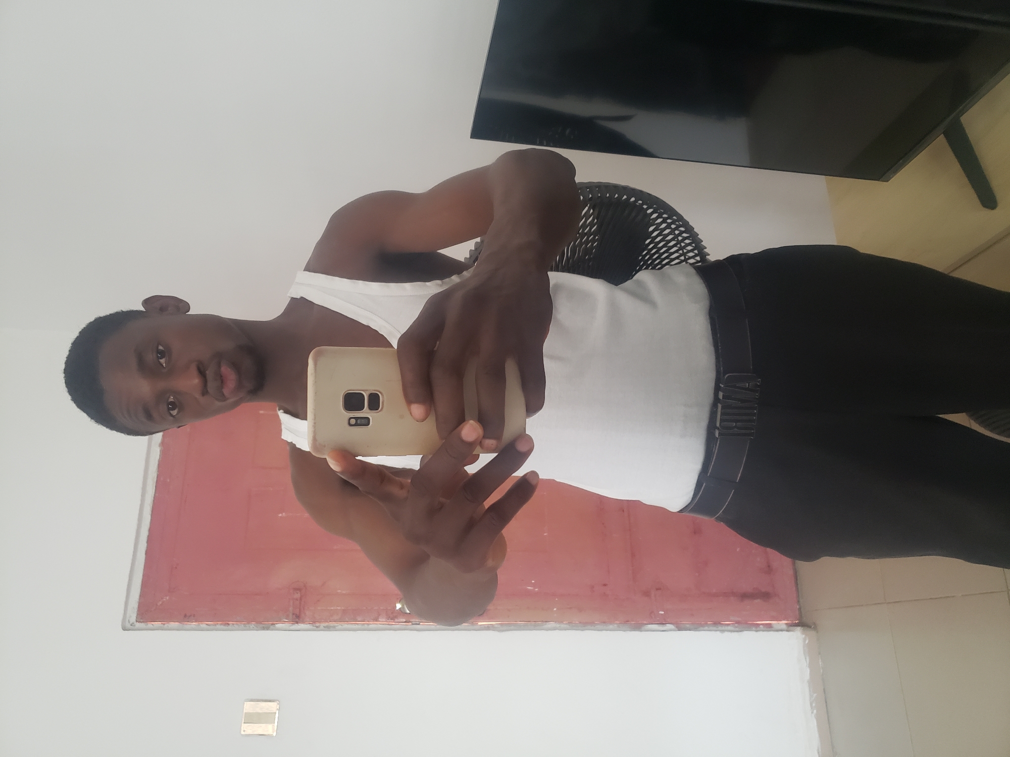 The image, presented rotated 90 degrees counter-clockwise, features a young adult male taking a mirror selfie in an indoor setting, likely in Adonkolo, Nigeria. The subject has dark skin, short dark hair, and is looking directly at the camera with a neutral expression and slightly parted lips. He is wearing a white tank top and black pants with a dark belt.
He holds a light-colored smartphone, identifiable by its triple camera array on the back, in his right hand to capture the image. His left hand is raised, with fingers spread in a gesture that resembles a peace sign.
The background elements suggest a domestic interior. A red-painted door frame is visible behind and to the man's left. Part of a black mesh chair is seen behind his right arm. In the upper right background, a black flat-screen television is present, possibly mounted above a light brown wooden surface. The walls are white, and the floor, visible in the bottom right, consists of light-colored tiles. A light switch or electrical outlet is also visible on the white wall. The scene is illuminated by ambient indoor light. Mirrored text, which appears to read "AMIRI," is faintly visible on his belt buckle.