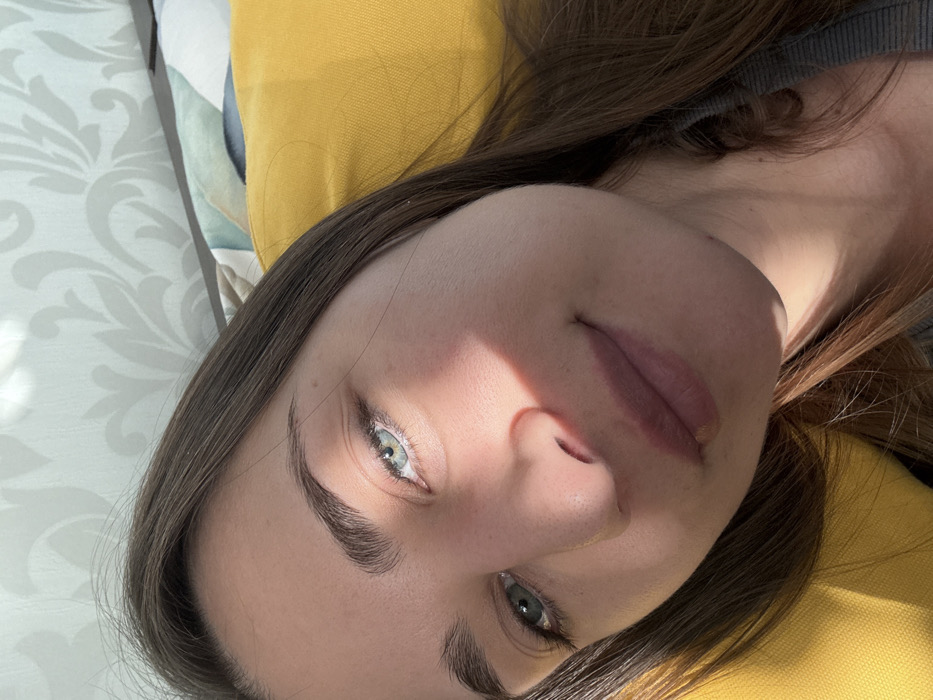 The image depicts a close-up, diagonal selfie of a young adult female in an indoor setting. The subject has fair skin, light blue-green eyes, dark eyebrows, and long, straight dark brown hair. She exhibits a subtle, closed-lip smile. Her attire includes a grey ribbed long-sleeved top, partially visible on the lower right. Strong sunlight illuminates the upper-left side of her face, casting shadows across her right cheek and chin, and highlighting her left eye and parts of her nose and forehead.
Her head rests against a bright yellow textured pillow. Behind this, a pillow with an abstract, multi-colored pattern in shades of green, blue, and white is partially visible. The background consists of a light-colored wall covered in wallpaper featuring a subtle, repeating floral or damask pattern. There are no identifiable landmarks, architectural features, or unique environmental cues within the image to determine a specific location within Saint Petersburg, Russia. The scene appears to be a residential interior.