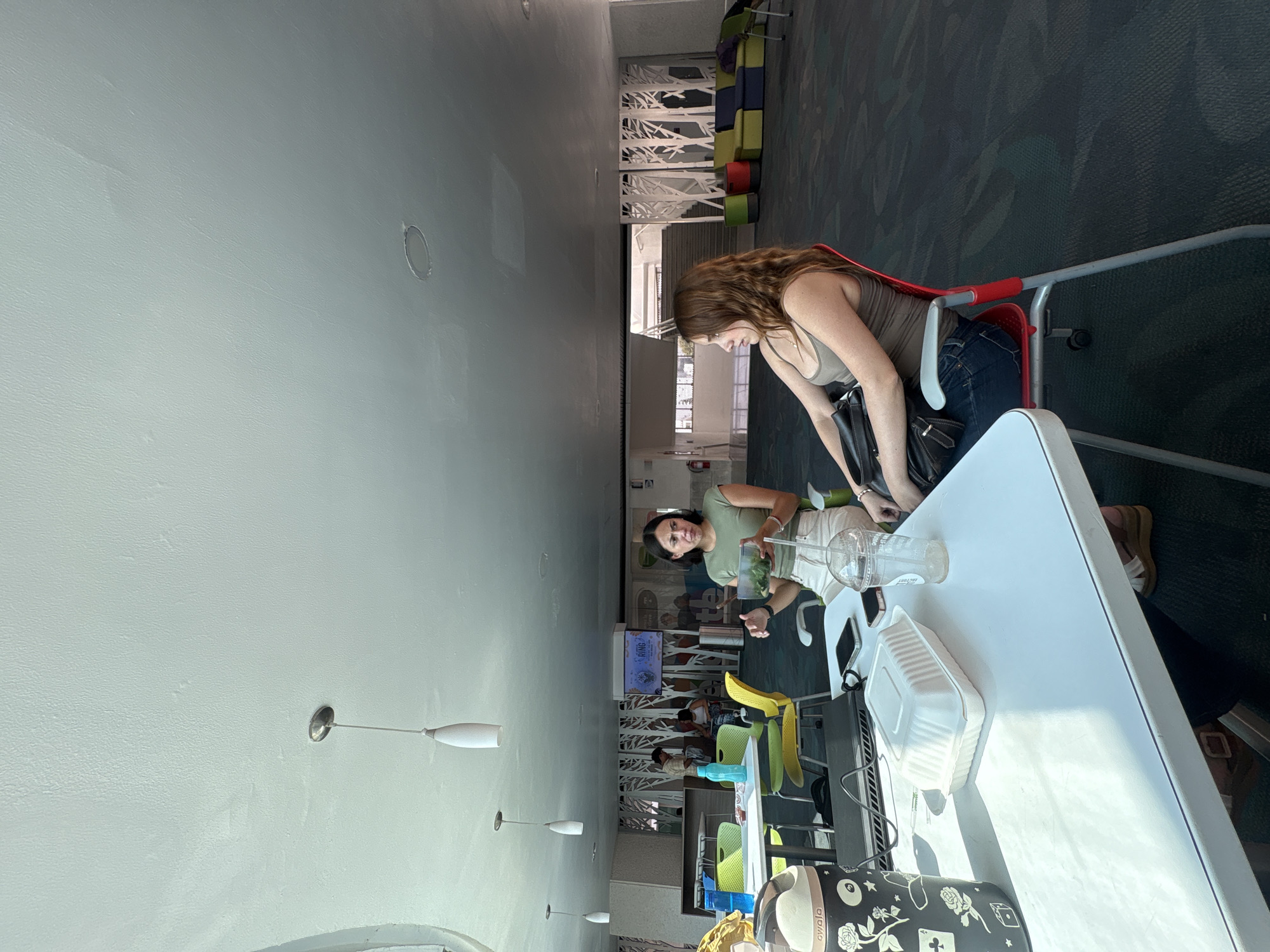 Two women are at a table in a brightly lit, modern-looking space, likely a library or cafeteria. One woman is sitting in a red chair, leaning towards the table, and is wearing a green tank top and blue jeans. The other woman is standing and looking at the contents on the table, wearing a green short-sleeved shirt and khaki pants. On the table are two plastic cups, a Styrofoam container, a black water bottle, and a cell phone. The space features modern white architectural details, hanging lights, and colorful chairs in the background. A television screen displays an image. Based on the design and architecture, it's likely a public or semi-public space. It's not possible to determine if the location is in the city of Puebla, Mexico, or confirm its exact location within a specific city or building based on the image.