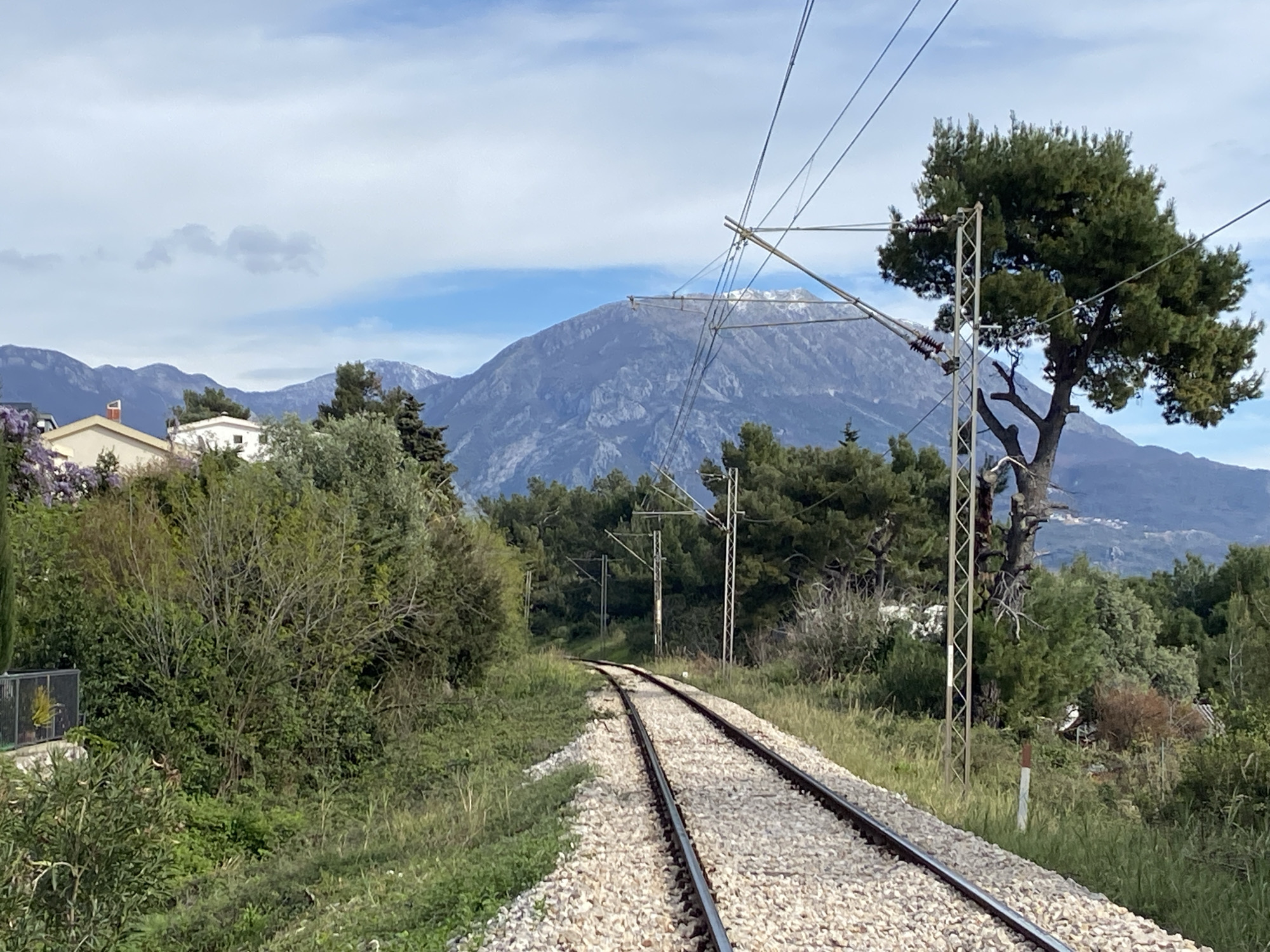 The image shows a section of a railway track curving through a green, vegetated area near Šušanj, Montenegro. The tracks are laid on a bed of light-colored gravel. Overhead power lines supported by metal poles run alongside the tracks. Lush greenery lines both sides of the railway, including various trees and bushes. In the background, a large mountain rises, its peak partially covered with light snow. A few houses are visible among the vegetation in the mid-ground, to the left. The sky is partly cloudy with patches of blue.