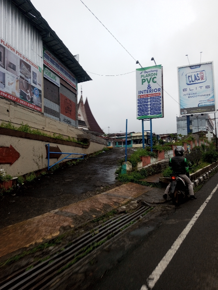 The image depicts an overcast street scene in Chagzam_Pam, Bhutan, characterized by a wet asphalt road and various commercial structures. On the far left, an elevated building with a corrugated metal facade displays signs advertising "PLAFON PVC & INTERIOR" and a business named "GEMERLANG," along with phone numbers. Below this building, a concrete slope leads upward, marked by a painted red arrow and a blue metal barrier. The ground here is uneven and damp, with scattered green vegetation.
In the mid-ground, two large billboards stand prominently. The left billboard, labeled "Gemerlang," advertises "PLAFON PVC & INTERIOR" with blue and green text. The right billboard promotes "NEW CLAS ELITE" with a white and blue design. Distant buildings with distinctive, steeply pitched roofs are visible behind the billboards and to the left of the main slope, suggesting traditional architecture.
On the right side of the main asphalt road, a single individual is seated on a dark motorcycle. The person wears a grey helmet, a dark vest with green shoulder accents, and light-colored pants. The motorcycle is stationary, facing away from the viewer. The road surface is dark and appears wet, featuring a linear drainage grate and a worn white lane marking. Sparse green foliage grows along the road's edge and on low retaining walls. Overhead, a single utility wire traverses the frame against the bright, overcast sky.