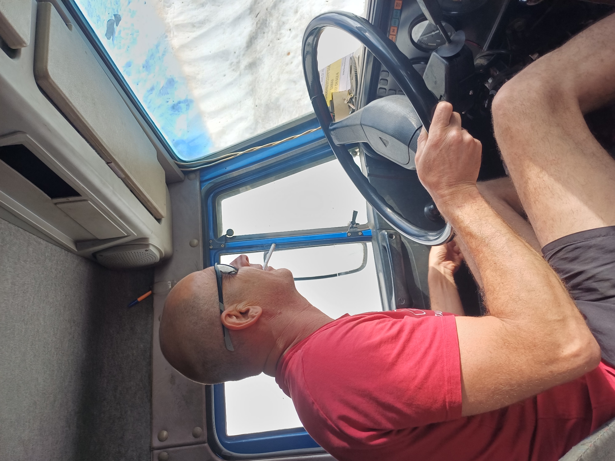 A man with a shaved head and glasses leans back in the driver's seat of a vehicle, holding a cigarette to his mouth. He is wearing a red t-shirt and appears to be looking upwards towards the sky or a window above. The interior of the vehicle is visible, including a steering wheel and dashboard. A yellow piece of paper with text is attached to the steering wheel. The scene is outdoors, with bright sunlight suggesting it is daytime. There is a hint of blue in the upper part of the image, possibly from the sky or reflections. A pen with an orange tip rests on a grey surface to the left of the man. The overall impression is one of a casual moment, perhaps during a break from driving.