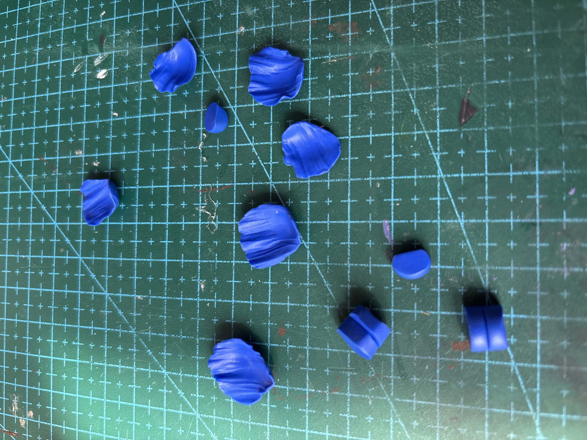 A close-up, overhead shot shows various small, curved, blue objects scattered across a green grid cutting mat. The objects appear to be made of clay or a similar pliable material and are molded into petal-like shapes, some with defined creases and edges. A few of the objects are cylindrical or semi-circular in form. The cutting mat is marked with a grid of turquoise lines, with intersecting horizontal and vertical lines and diagonal lines. Some faint smudges and red marks are visible on the mat. There are no people or animals in the image, and the scene appears to be a tabletop workspace. The image does not provide any information about the location in Blagoveshchensk, Russia.