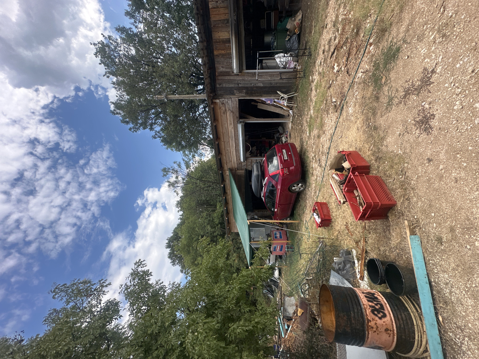 A wide shot captures an outdoor scene featuring a red car parked in front of a wooden structure that appears to be a garage or workshop. The structure has two open bays, one with a red car and the other with various items including bags and a rack. Red plastic crates, some filled with debris, are scattered on the ground in front of the garage. To the left of the garage, a large, rusty barrel with "OLMA" written on it stands near other discarded materials and a green net. Trees and foliage surround the area, and the sky above is blue with scattered white clouds. The ground is a mixture of dirt and gravel. The location is identified as Cetinje, Montenegro. There are no people visible in the image.