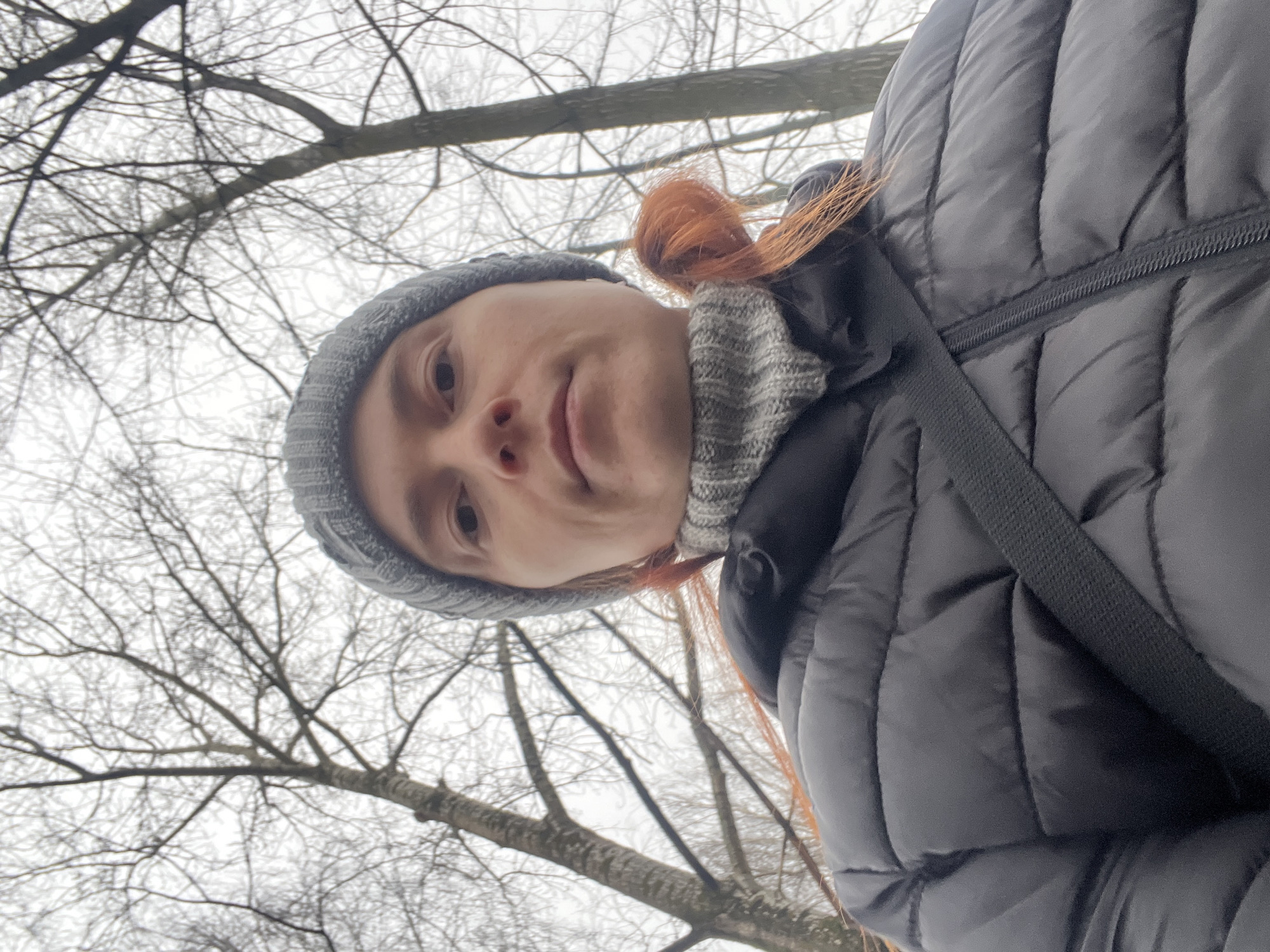 A low-angle selfie captures a person wearing a gray knit hat and a dark puffy jacket, with orange hair visible. A gray knitted scarf is also visible around the neck. The person's face is centrally framed, showing facial features like eyes, nose, and lips. The background reveals bare tree branches against a pale, overcast sky.
There is not enough information to determine the specific location in the city of Chaikovsky, Russia.