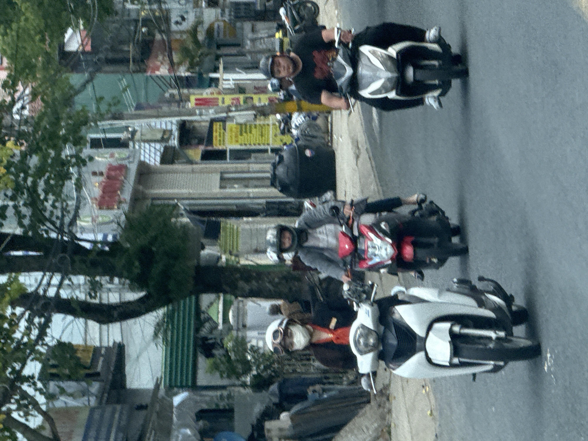 The image captures a street scene in Lâm Viên - Đà Lạt, Vietnam, with three individuals on motorcycles. The person in the foreground on the left is a woman wearing a white helmet with goggles and a white face mask, a red shirt, and a dark jacket. She is riding a white scooter. To her right, a person in the middle, wearing a dark helmet and a grey jacket over a lighter shirt, rides a red and black scooter. The person on the right, a man with a dark baseball cap and a black t-shirt with a graphic design, is on a white and black scooter. The environment is a paved street with buildings lining the sides, including structures with visible signage. Trees with green foliage are present along the street, and electrical wires are visible overhead. The overall impression is a typical urban street environment.