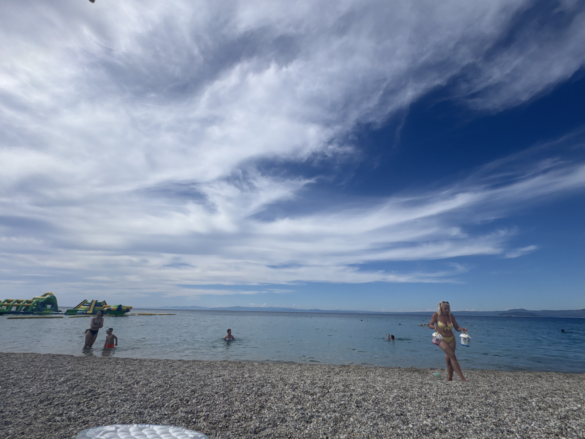 In Pefkochori, Greece, a wide, pebbled beach meets the clear blue water of the Aegean Sea under a bright, partially cloudy sky. The water is calm, with several people visible. In the foreground, on the beach, a blonde woman in a yellow bikini walks along the shore, carrying two buckets, one red and one white, suggesting she is heading for or coming from a beach activity. Further in the water, a woman in a black bikini and a young boy in orange swim trunks stand near the shore, with other people scattered in the distance. To the left, a large inflatable water park with slides and obstacles is partially submerged in the sea. The horizon shows a faint line of hills or mountains, indicating the presence of distant land. The overall scene captures a typical summer day at a Mediterranean beach.