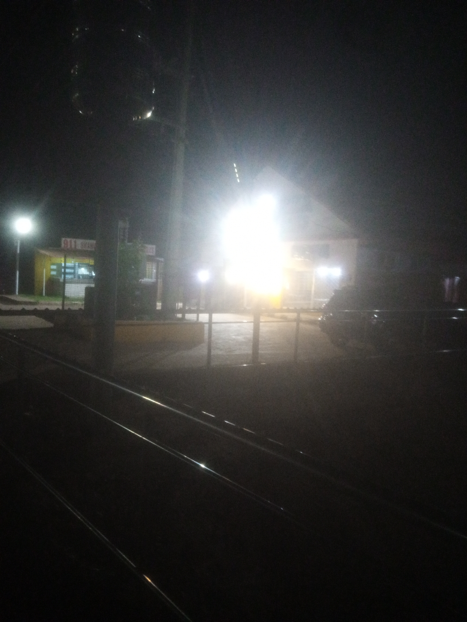 The image depicts a dimly lit outdoor scene at night, likely in the vicinity of a railway track, with a small structure resembling a shop or food stall in the background. A bright light source, possibly a streetlamp or floodlight, illuminates the scene, creating strong glare and obscuring details. The shop has a yellow facade with some lettering that is unreadable due to the lighting conditions. Several poles are visible, one of which appears to be a utility pole supporting wires. In the foreground, two parallel railway tracks are present, reflecting the ambient light. A dark, indistinct vehicle is parked to the right of the shop. The overall impression is that of a somewhat rural or semi-urban setting at night, characterized by low light and stark illumination. The specific location of Television, Nigeria, is not visually identifiable from the image itself.