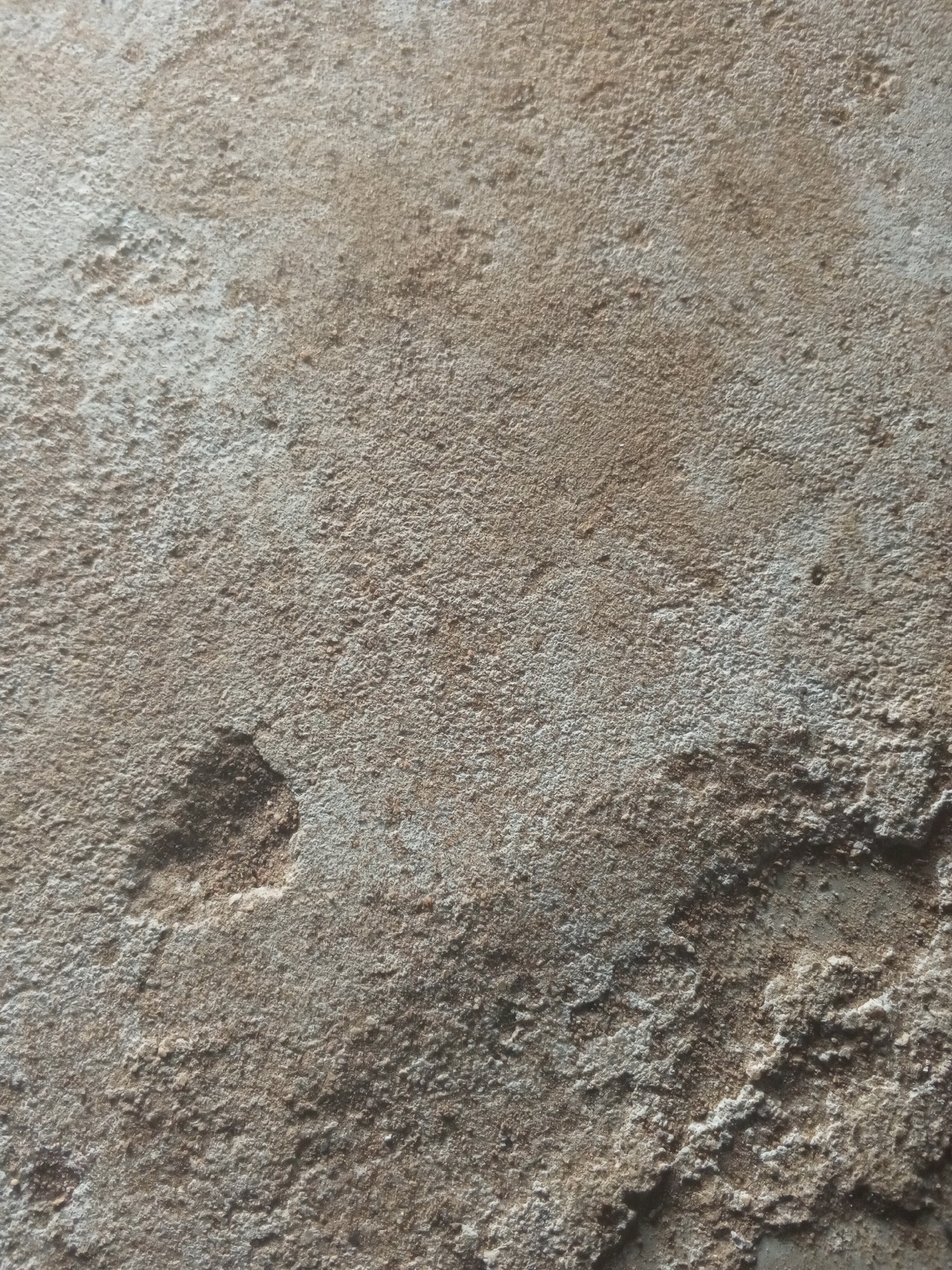 The image shows a close-up view of a rough, textured surface, possibly concrete or a similar building material. The surface is uneven with many small bumps and indentations. The color palette consists of various shades of brown, gray, and white, suggesting a weathered or unpainted material. There is a darker, irregularly shaped depression in the lower left quadrant of the image, which might be a chip or a natural imperfection in the material. The lighting creates shadows that emphasize the texture and contours of the surface. There are no people, animals, or discernible objects in the image, only the abstract texture of the material itself. The context of Nyabe, Nigeria, does not provide any specific visual cues within the image itself, as it solely focuses on the textural detail of the surface.