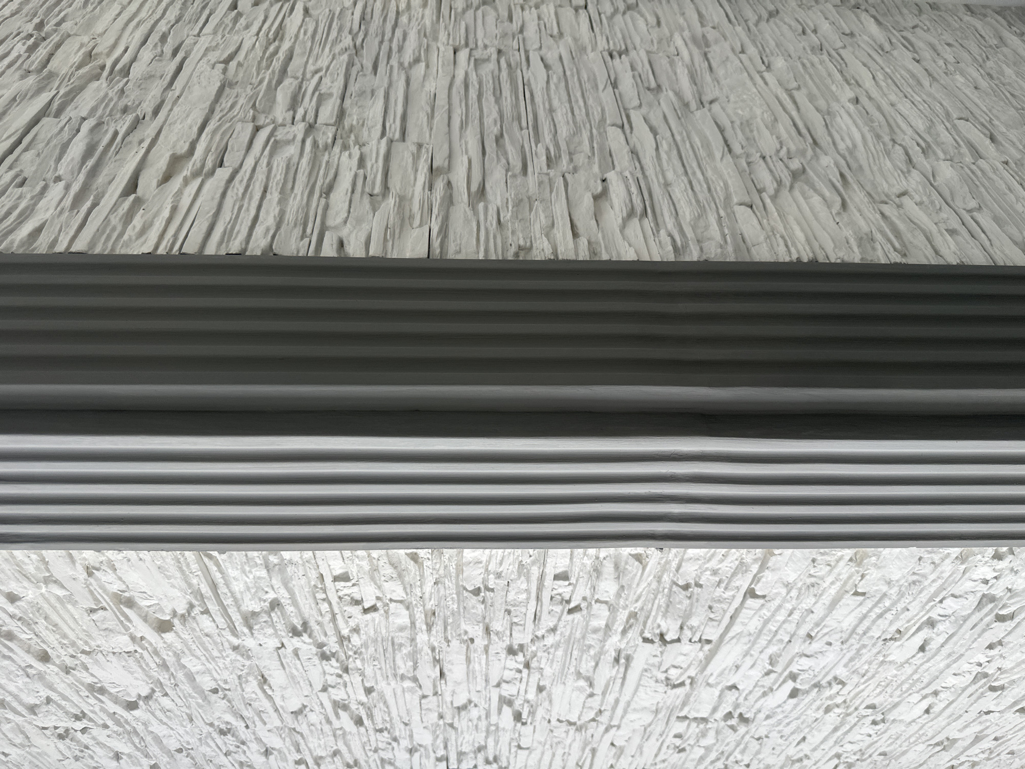 The image shows a textured white wall with a gray horizontal band in the middle. The wall texture is rough and jagged, resembling stacked stones or uneven plaster. The gray band features a series of parallel lines or grooves. The location is unspecified.