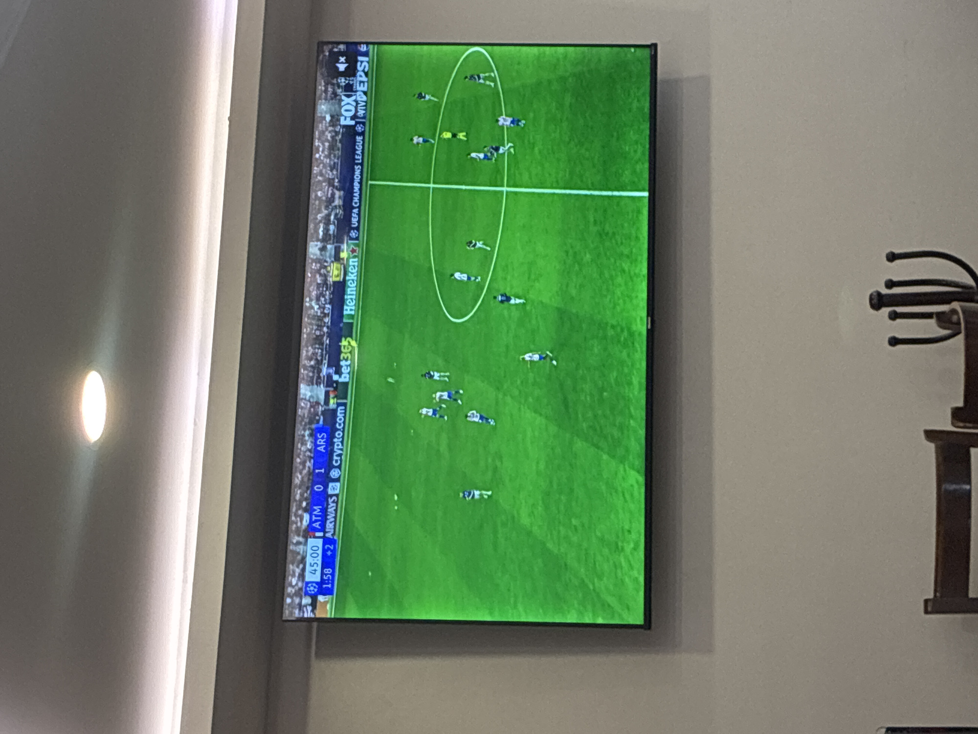 The image captures an indoor scene in Cuautitlán Izcalli, Mexico, featuring a wall-mounted television displaying a football (soccer) match. The television screen dominates the central part of the frame, showing a vibrant green football pitch with players in blue and white uniforms. A white oval marking is visible on the field.
Overlay text on the TV screen indicates it's a "UEFA CHAMPIONS LEAGUE" match broadcast on "FOX." Various sponsor logos are also visible, including "Heineken," "bet365," "crypto.com," "AIRWAYS," and "PEPSI." The scoreboard displays "45:00 ATM 0 1 ARS," with an additional "1:58 +2" below it. This suggests that Atlético Madrid (ATM) is losing 0-1 to Arsenal (ARS) at the 45-minute mark, with two minutes of added time.
To the left of the television, a circular, illuminated wall light fixture casts a warm glow on the light-colored wall. To the right, a dark, multi-pronged decorative item or coat rack is partially visible against the same wall. The overall setting suggests a casual viewing environment, likely a living room or a common area. The time shown on the screen implies the match is nearing the end of its first half.